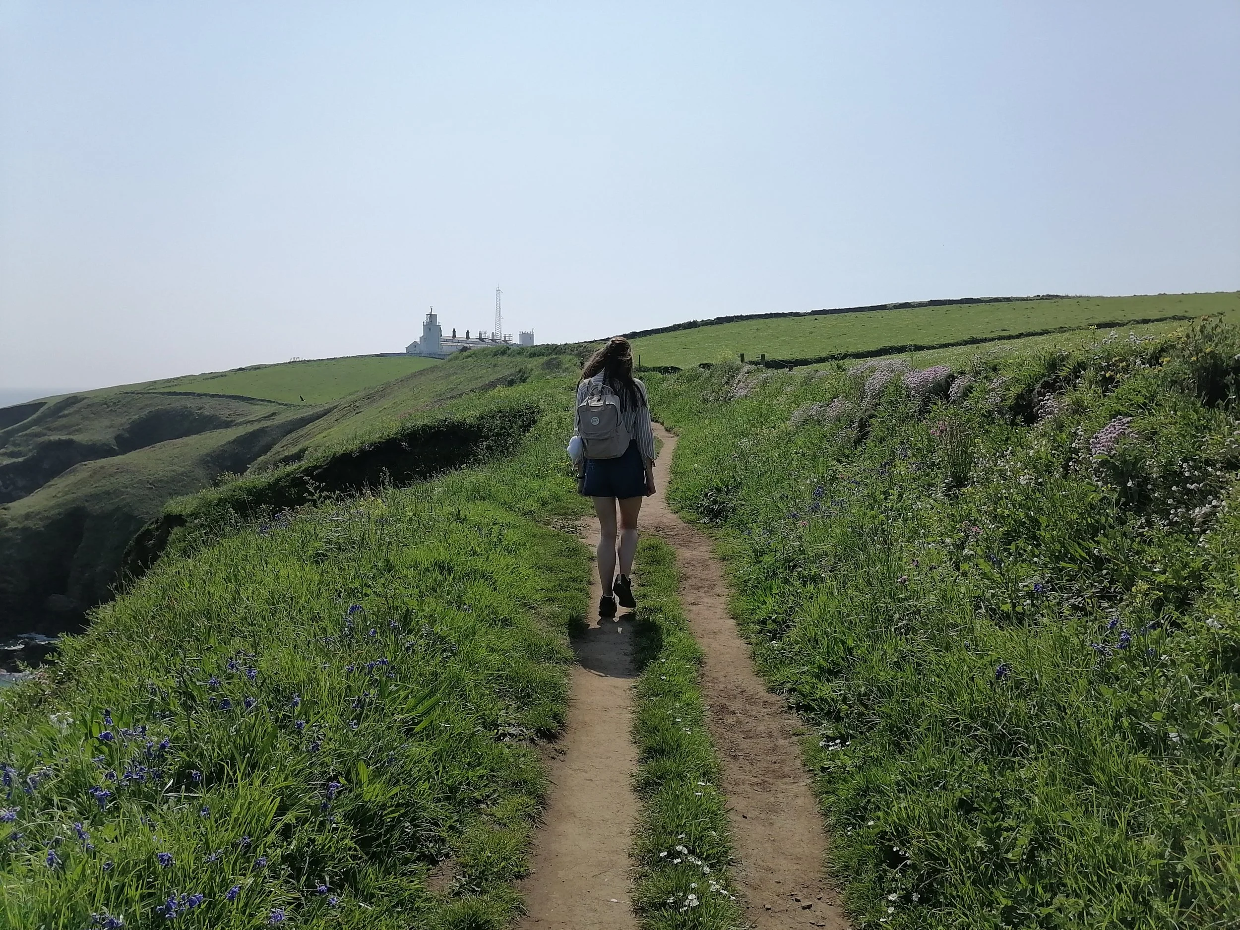 A woman with long hair, wearing a backpack, walking along a dirt trail through green hills with vibrant wildflowers, towards a lighthouse in the distance, under a clear blue sky.