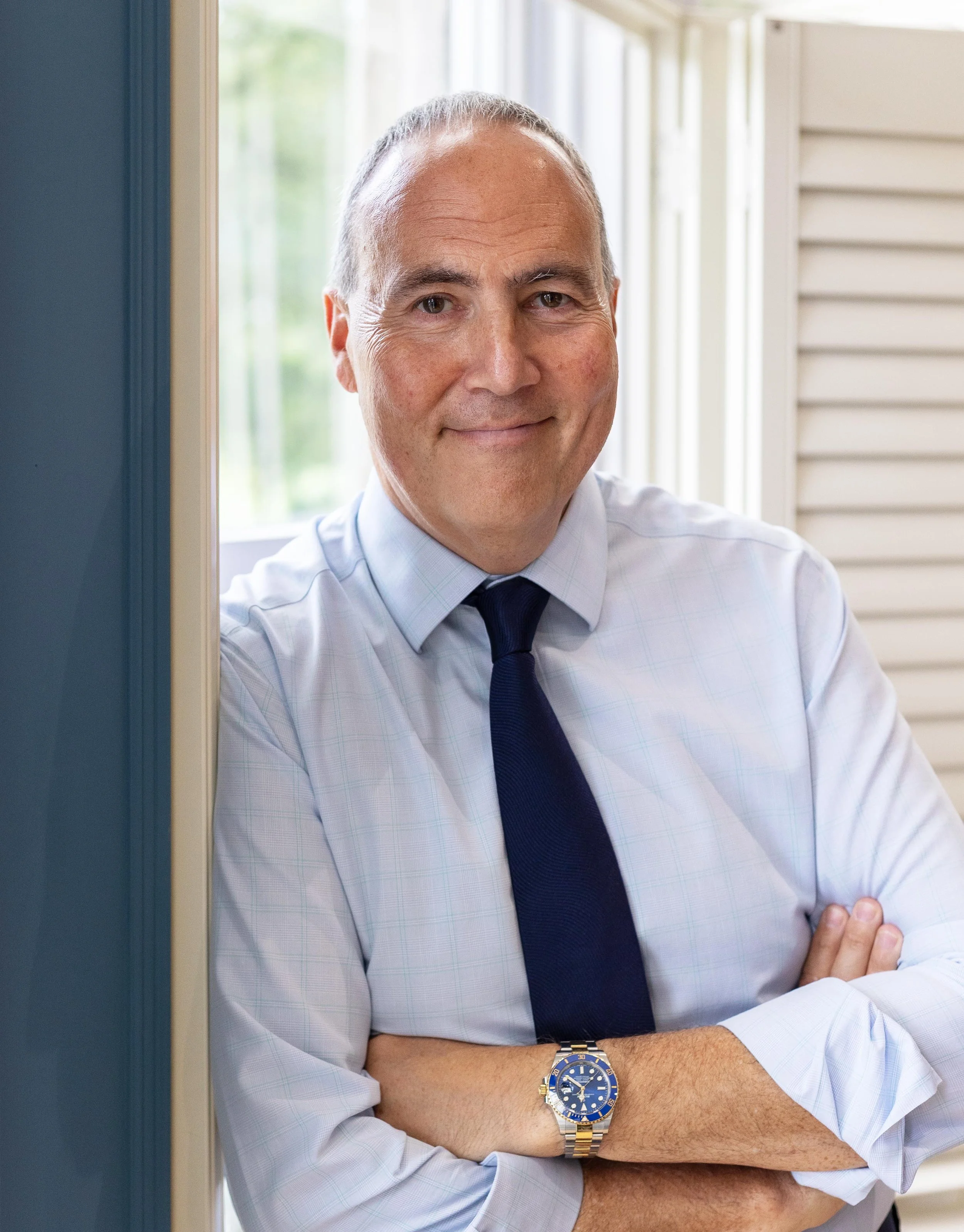 A middle-aged man with gray hair and light skin, wearing a light blue dress shirt, dark blue tie, and a wristwatch, standing with arms crossed at the front door of a house with white siding and a window, smiling.