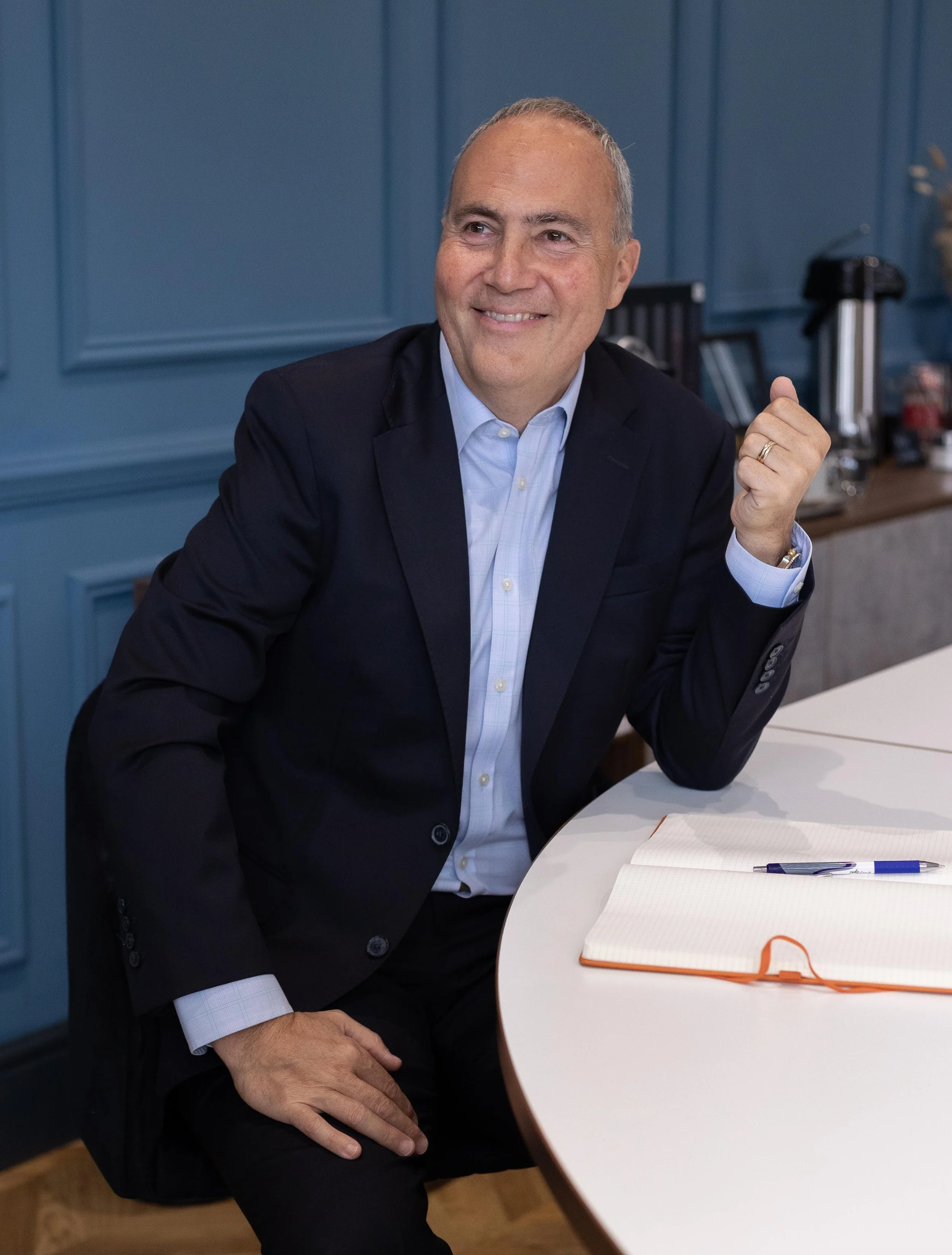 A man in a dark suit and light blue shirt smiling and sitting at a table with an open notebook and a pen in front of him, in a room with blue painted wood panel walls.