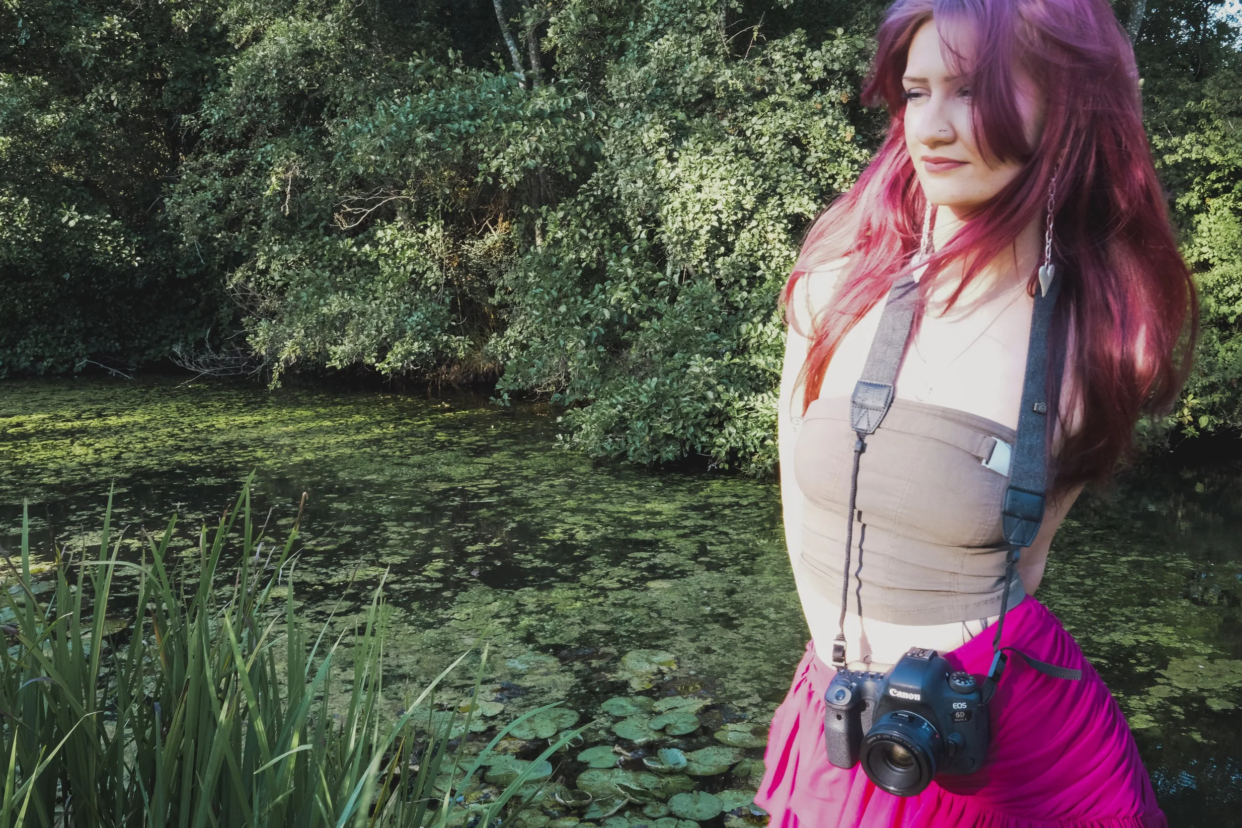 A woman with pinkish hair wearing a gray top and pink pants, carrying a camera around her neck, standing near a body of water covered with green duckweed, surrounded by dense green foliage.