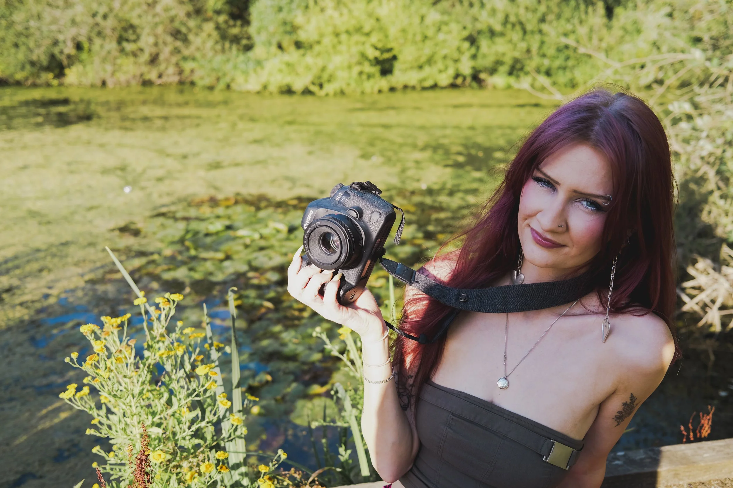 A woman with red hair, wearing a necklace and dark top, holding a camera, standing outdoors near a water body with greenery and yellow flowers.