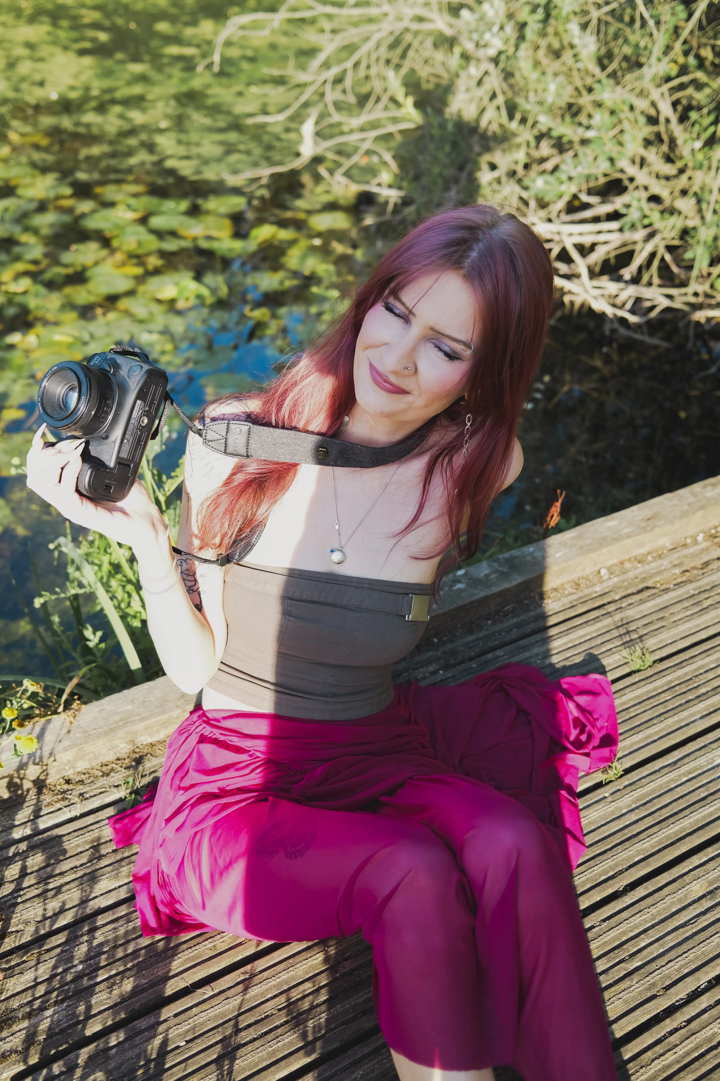 A woman with reddish-brown hair sitting on a wooden dock near water with lily pads. She is holding a camera and smiling with her eyes closed, wearing a beige top, bright pink pants, and jewelry.