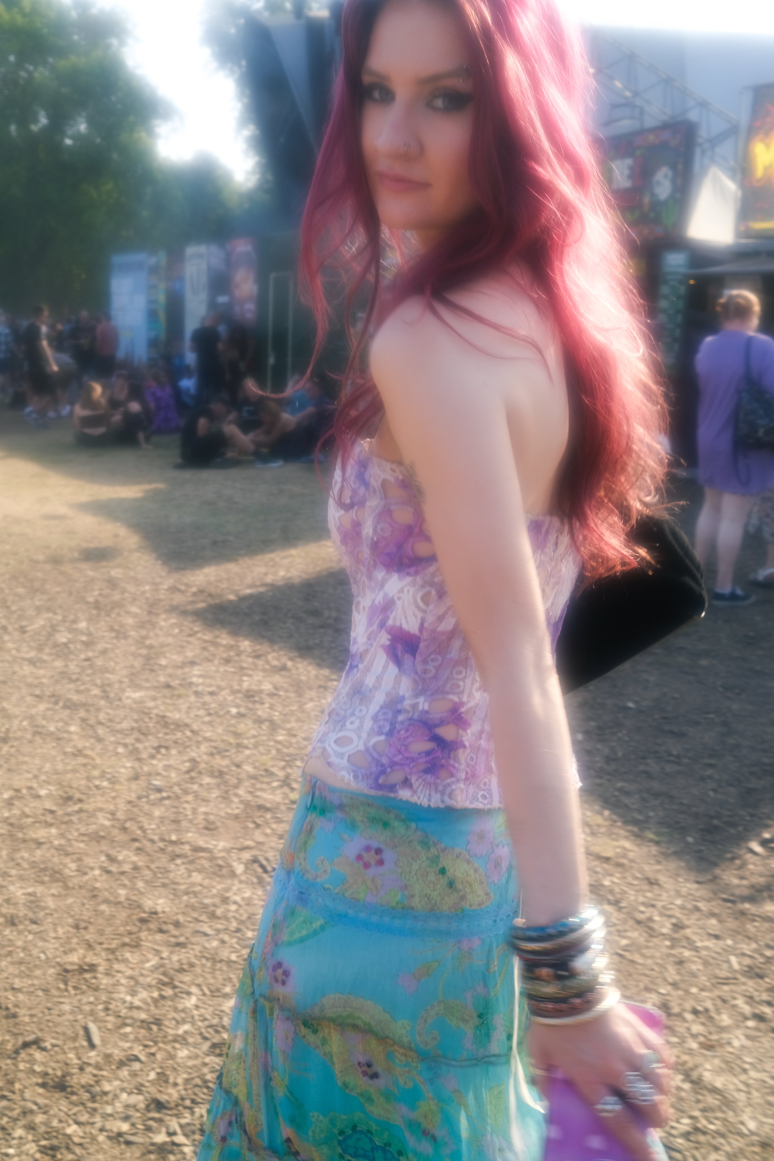 A young woman with pink hair wearing a colorful top and a long, patterned skirt, standing outdoors at a music festival with a crowd in the background