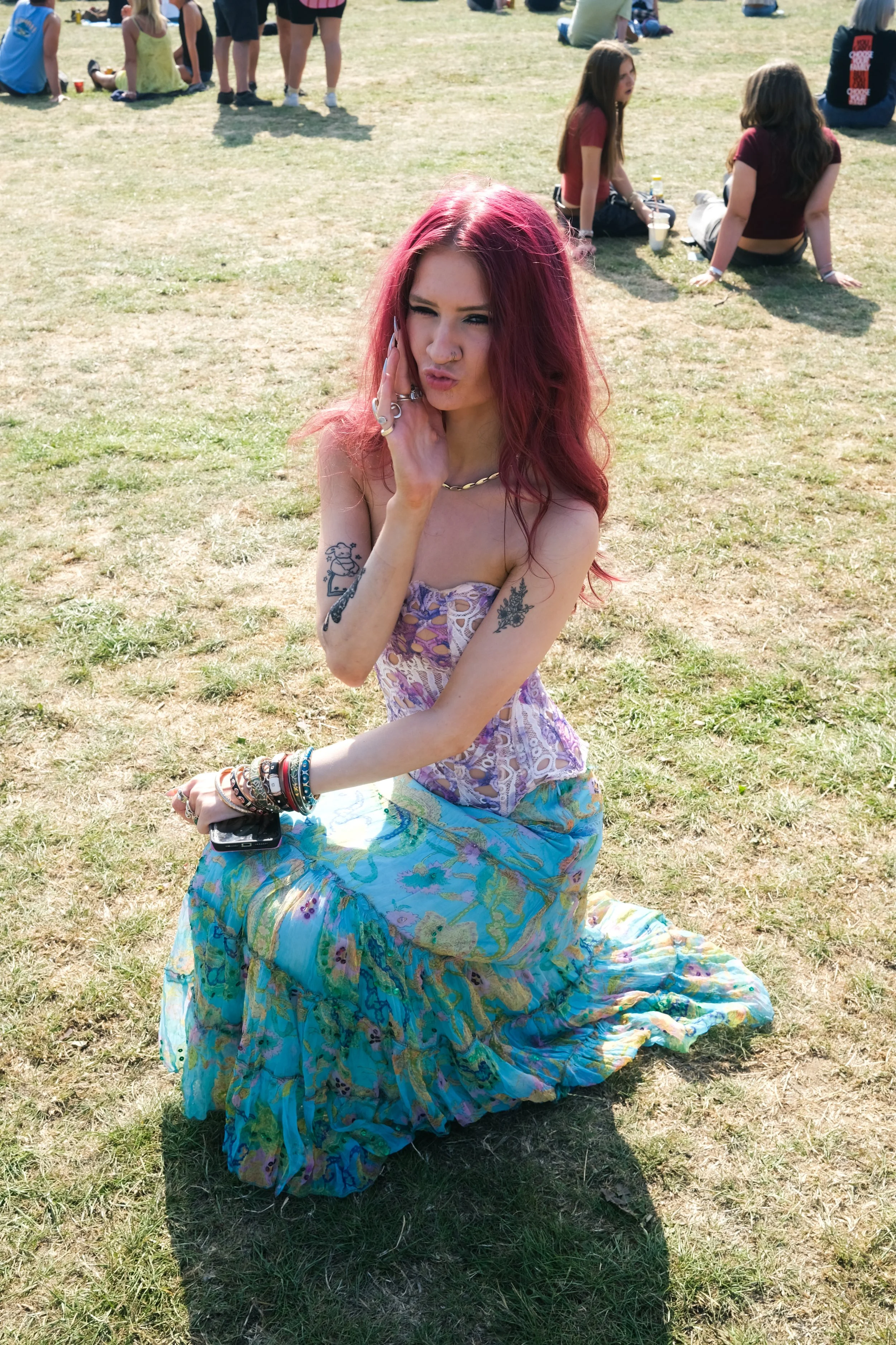 A woman with pink hair, tattoos, and layered bracelets, wearing a colorful patterned skirt and a strapless top, sitting on the grass at an outdoor event with other people in the background.