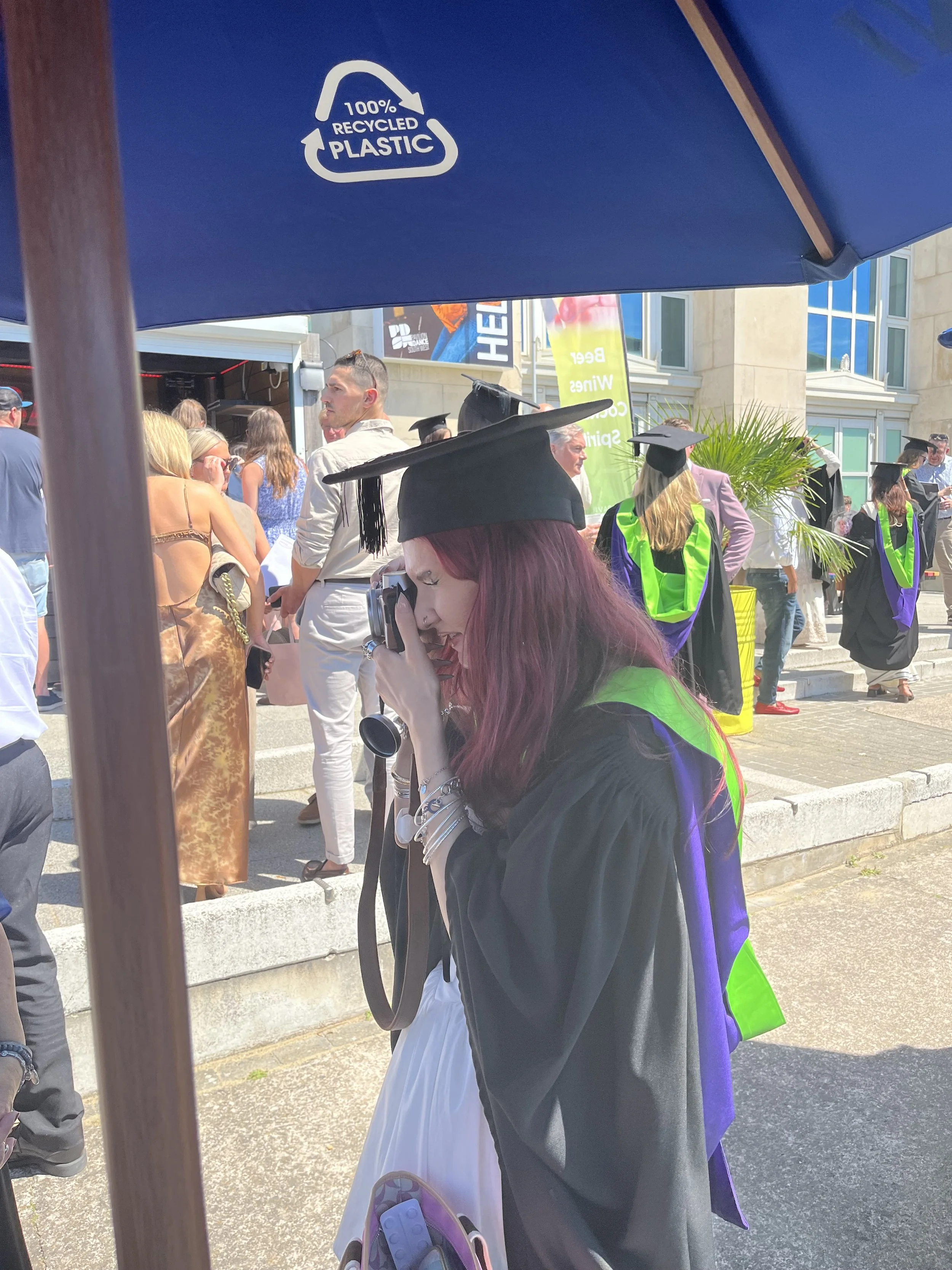 Group of people, including graduates in caps and gowns, gathered outdoors, with some wearing casual clothing and others in academic regalia, under sunny weather in front of a building.
