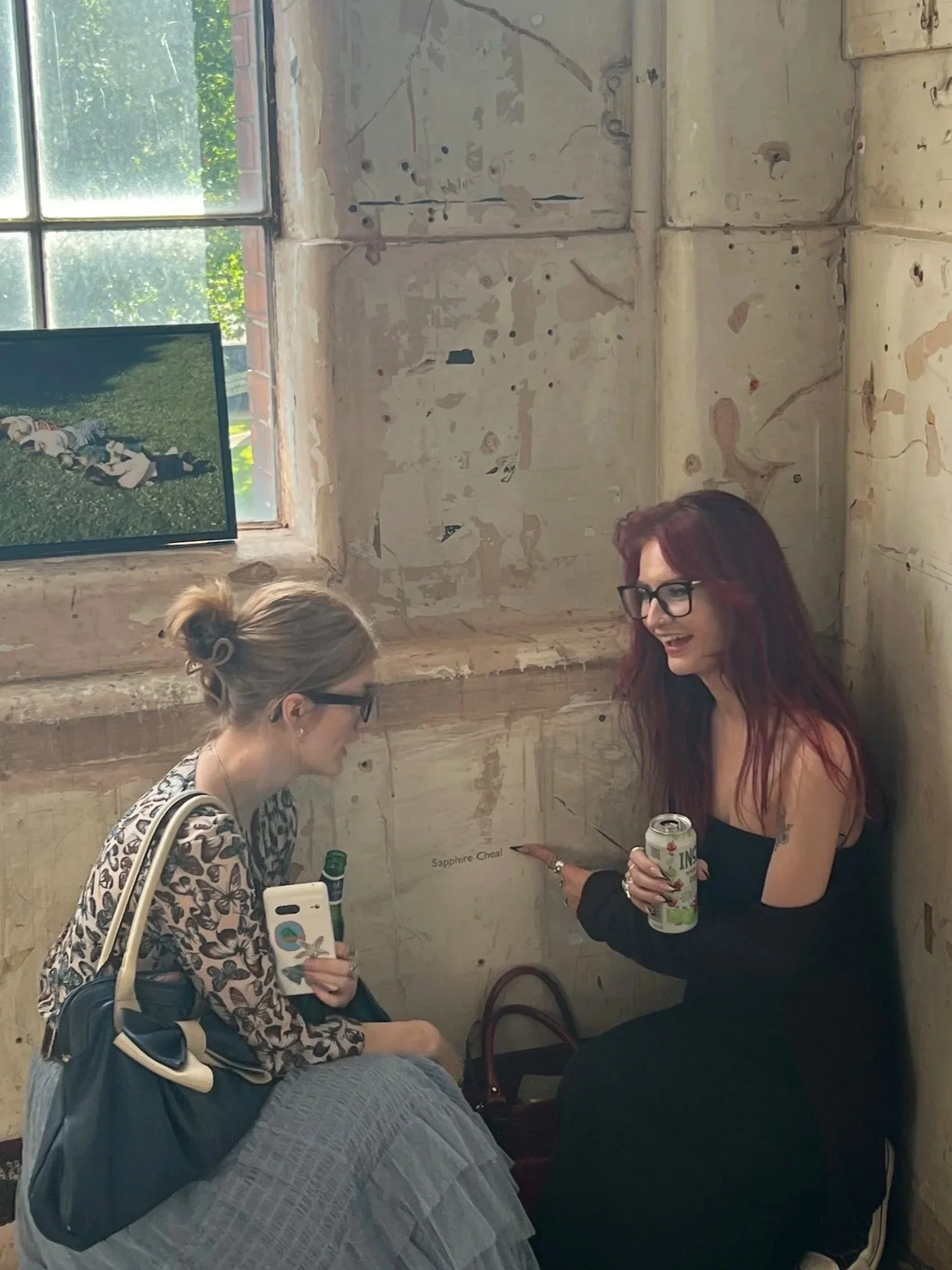 Two women are sitting inside a rustic, unfinished room with exposed wood and plaster walls. One woman with a high bun and glasses is holding a phone, the other woman with long reddish hair and glasses is smiling and holding a can of drink. A framed picture of a football game is hanging on the wall. Outside the window, trees and a brick wall are visible.