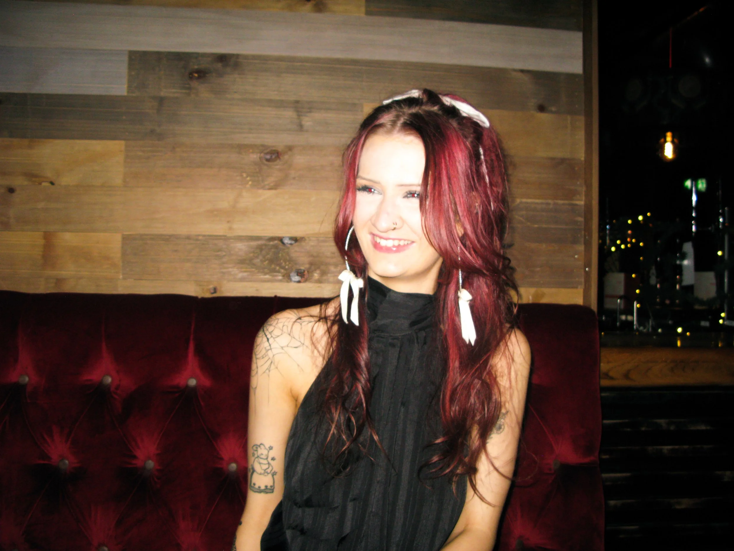 A young woman with long red hair, tattoos on her arm, and hoop earrings smiling in front of a wooden wall at night.