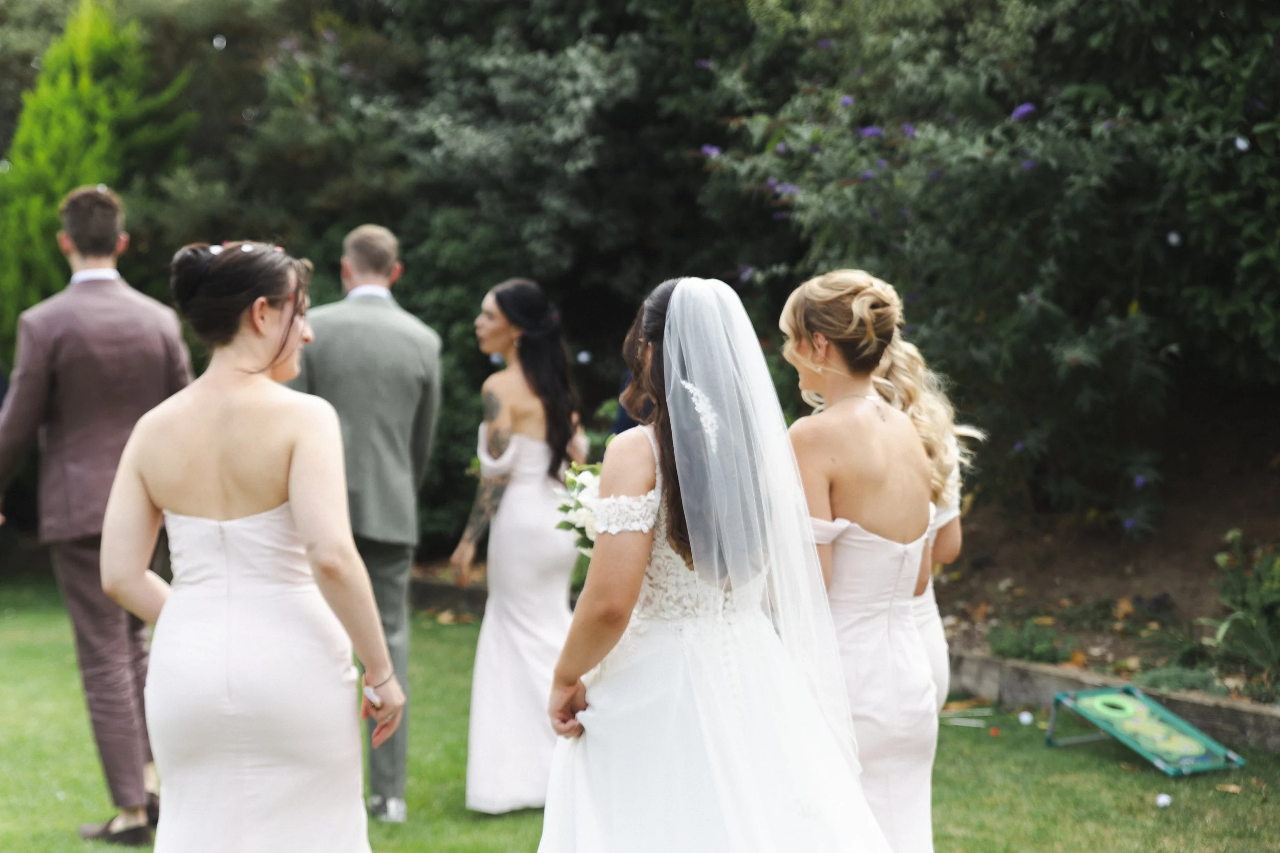 A group of people gathered outdoors in a garden, with some dressed in formal attire including a woman in a white wedding dress and veil, indicating a wedding ceremony.