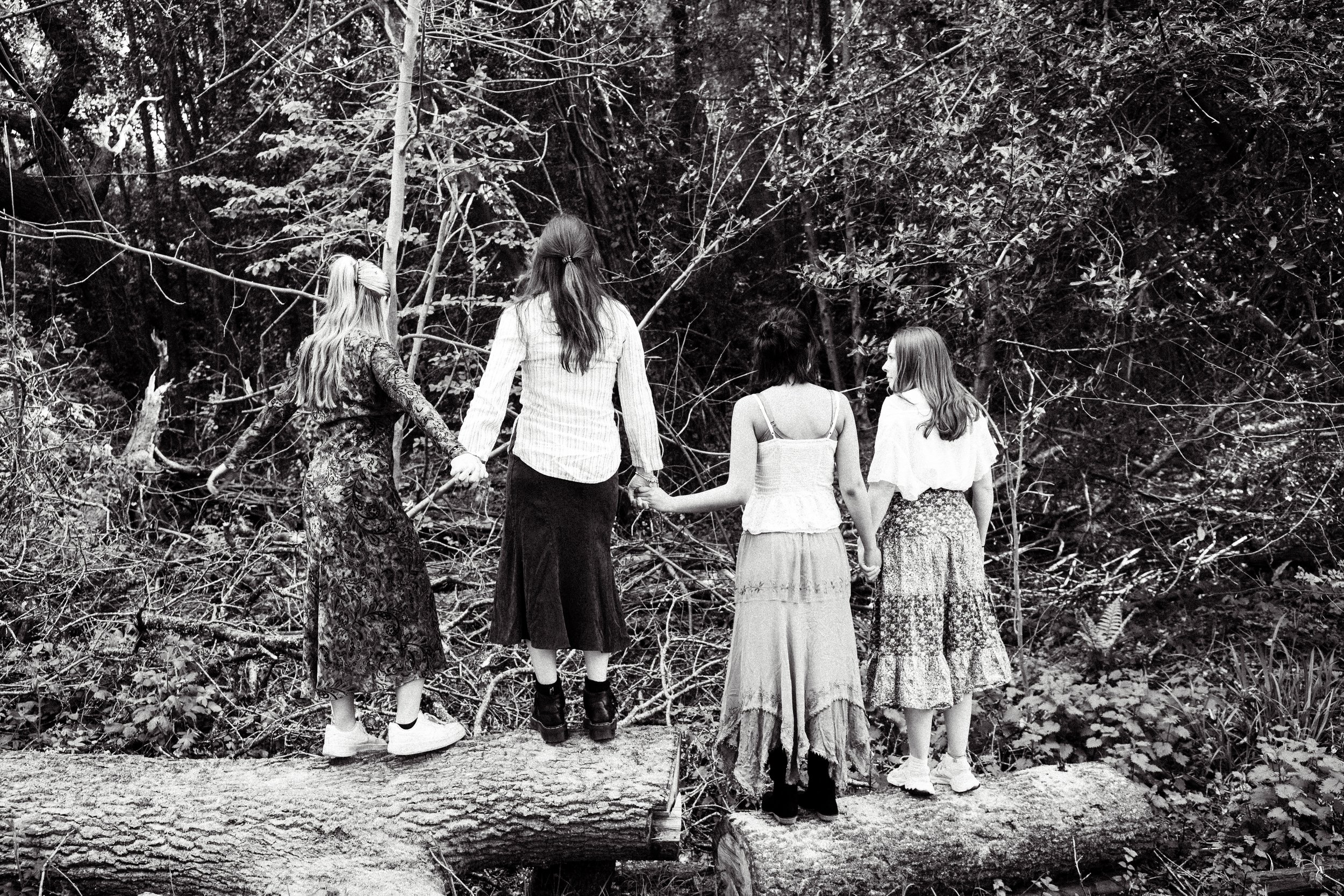 Four women standing hand-in-hand in a forest, facing away from the camera. The photo is in black and white.