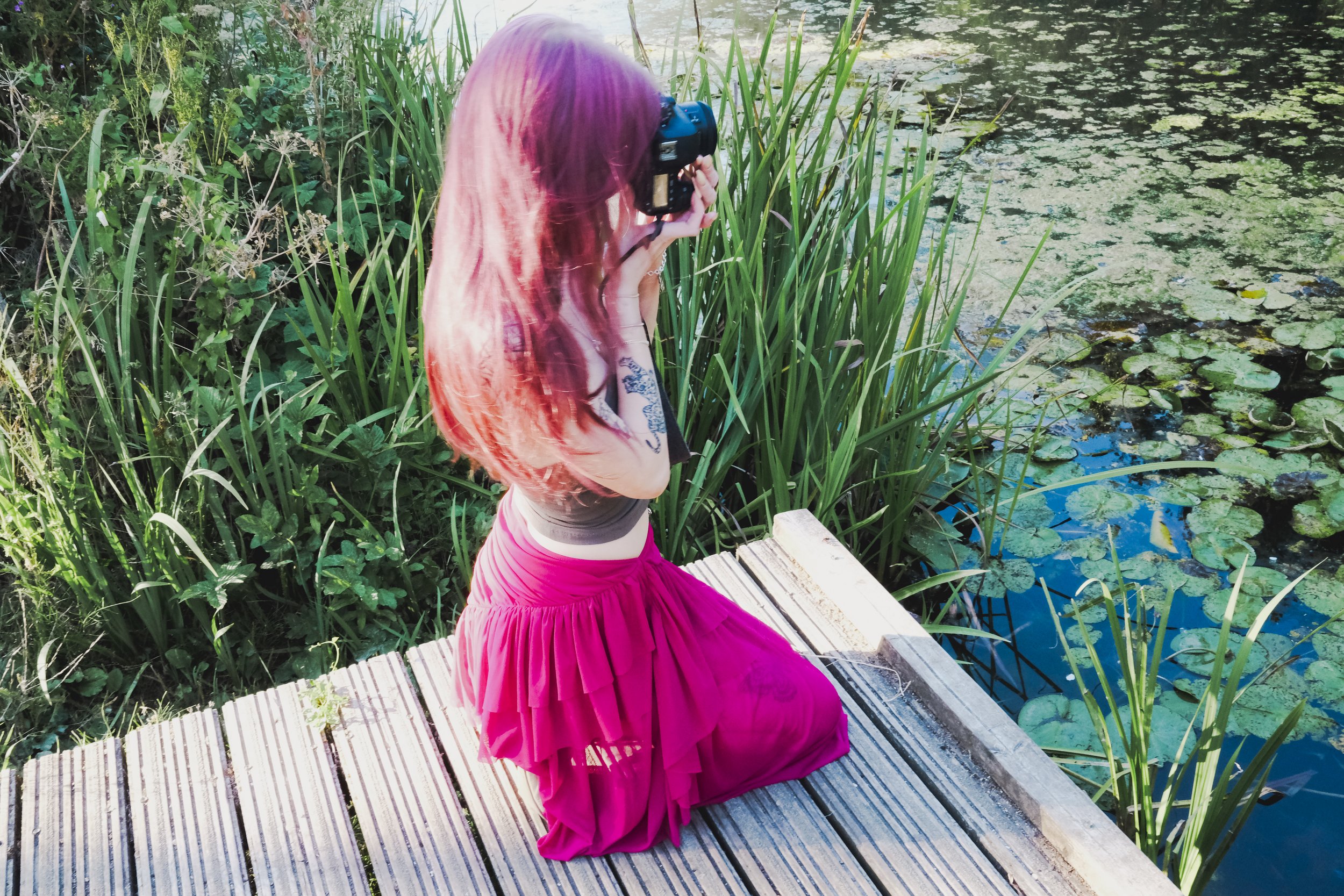 A woman with long red hair, dressed in a pink skirt and beige top, sitting on a wooden dock by a pond, taking a photograph of the water with a camera.