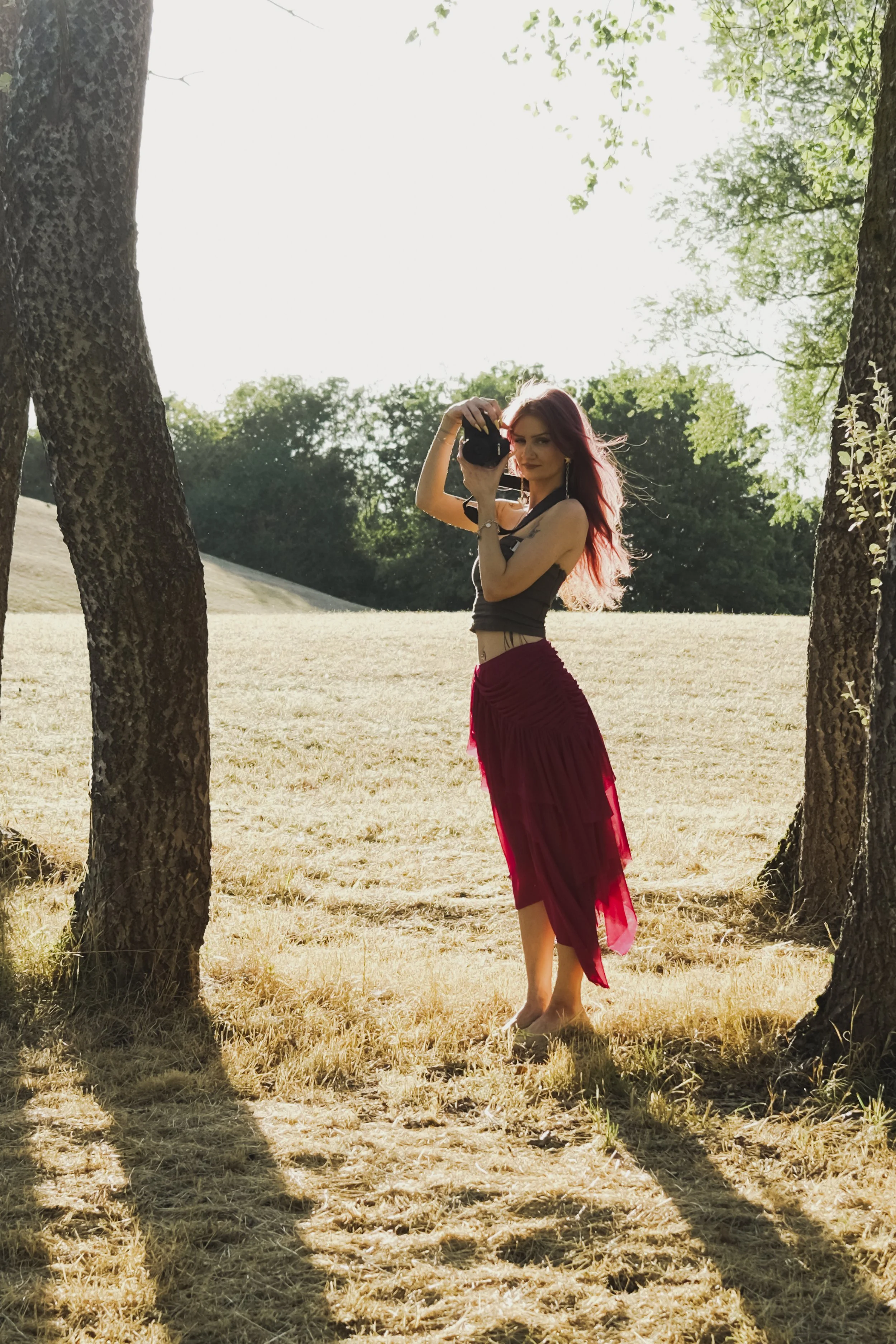 A woman with red hair and tattoos is standing outdoors between two trees, taking a photograph with a camera. She is wearing a black top and a long, red skirt, and is barefoot. The background features a grassy area, trees, and bright sunlight.