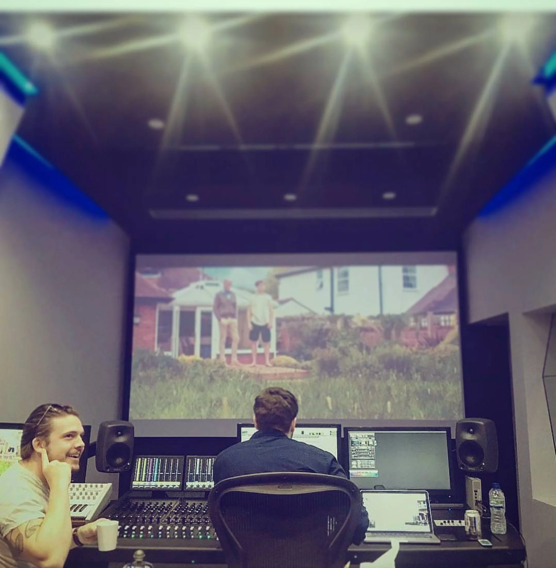 Recording studio with three people working at mixing consoles, one man facing away and two people in front, a large screen displaying a photo of two men in front of a house.