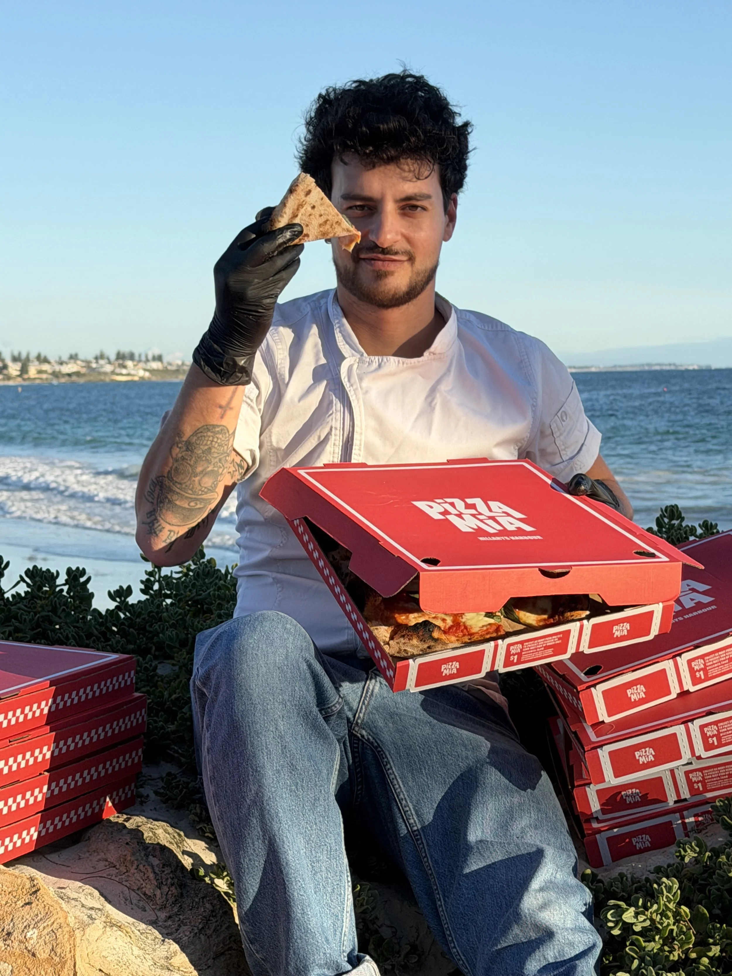 A man sitting on rocks near the ocean, holding a slice of pizza and surrounded by stacked pizza boxes. He has curly hair, tattoos, and is wearing a white shirt and jeans.