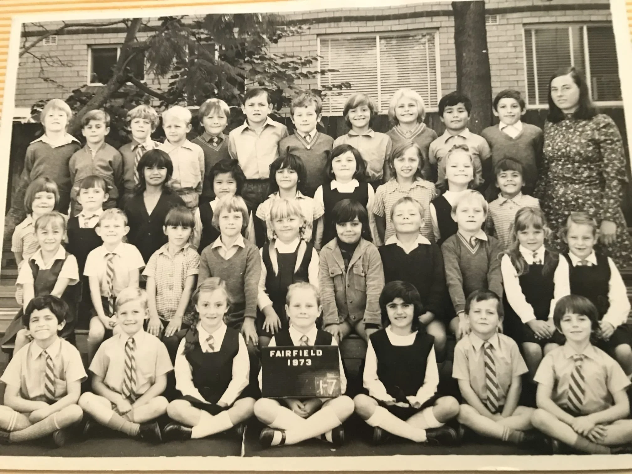 Black-and-white class photo of young students and their teacher outside school, with a sign reading 'Fairfield 1973', in front of trees and a brick building.