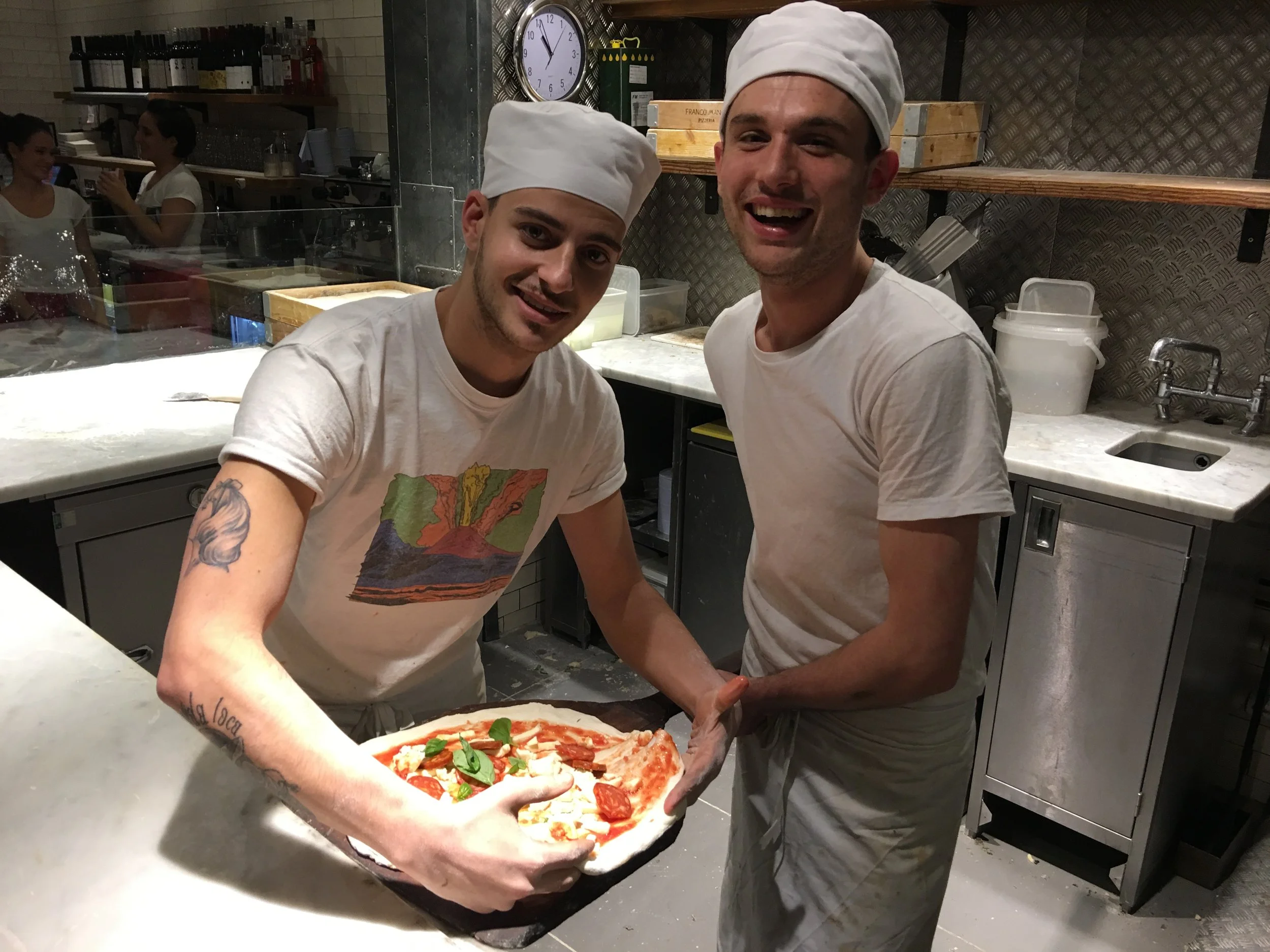Two male chefs wearing white chef hats and shirts smiling in a professional kitchen, holding a pizza topped with basil and other ingredients.