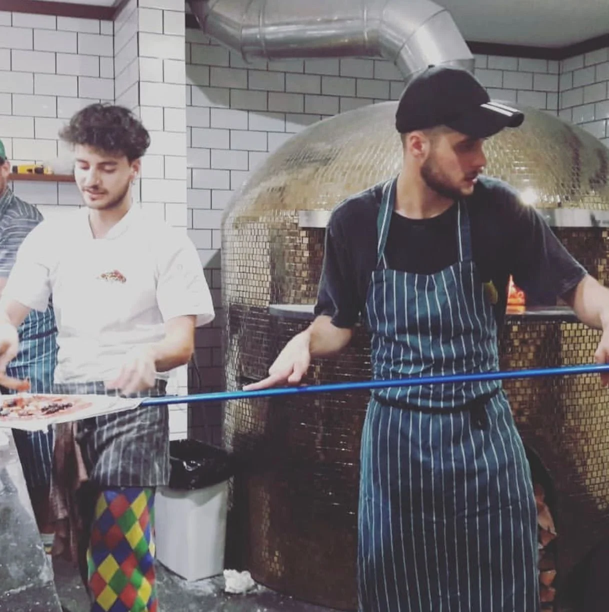 Two young men in striped aprons working with a pizza oven in a restaurant kitchen with white subway tile walls.