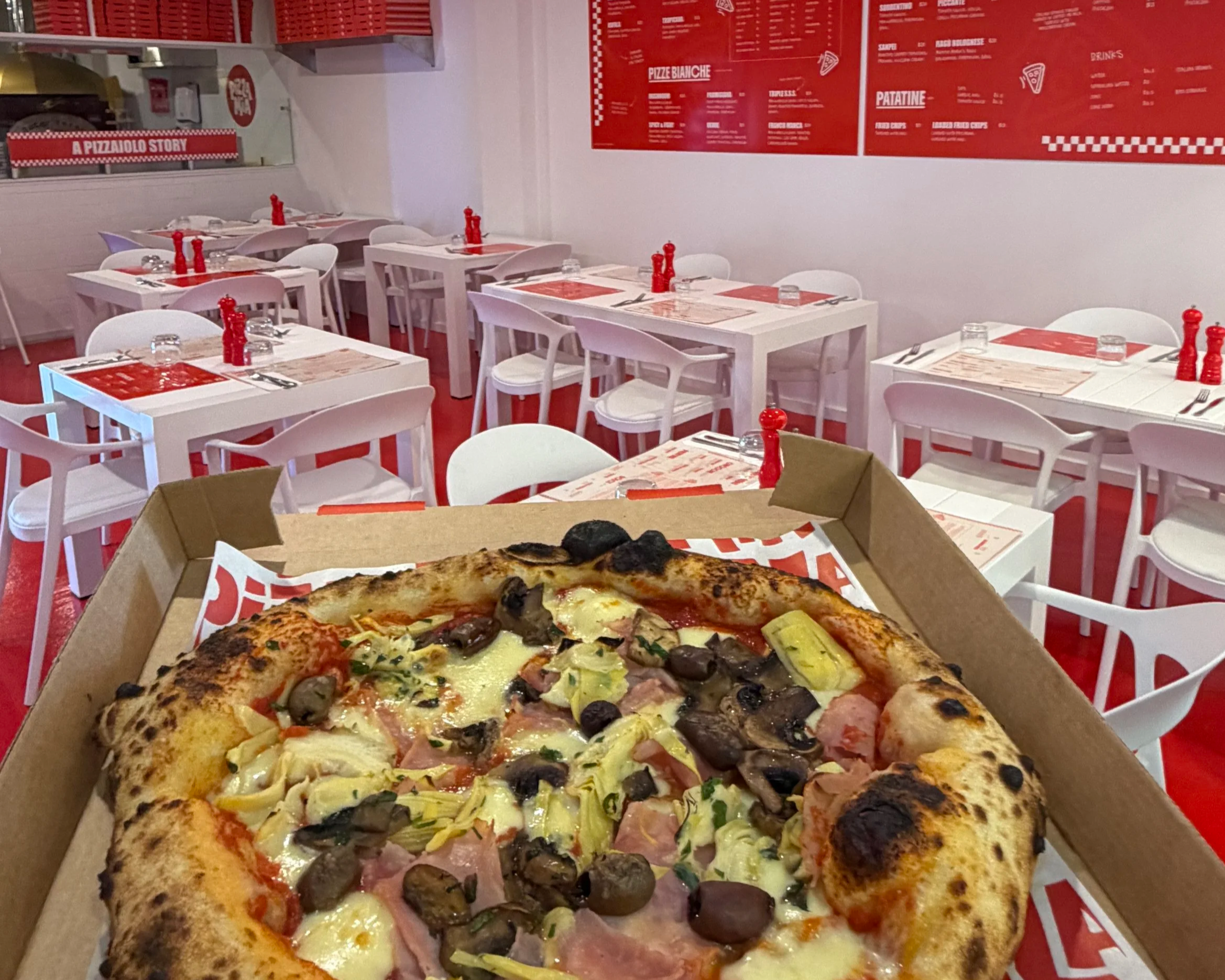 A pepperoni pizza with black olives, green peppers, and ham in a pizza box in the foreground, with a restaurant interior featuring white tables, chairs, and red wall menus in the background.