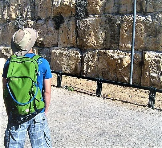 A young boy with a green backpack and a tan hat stands near a large rock wall, looking away from the camera on a sunny day.