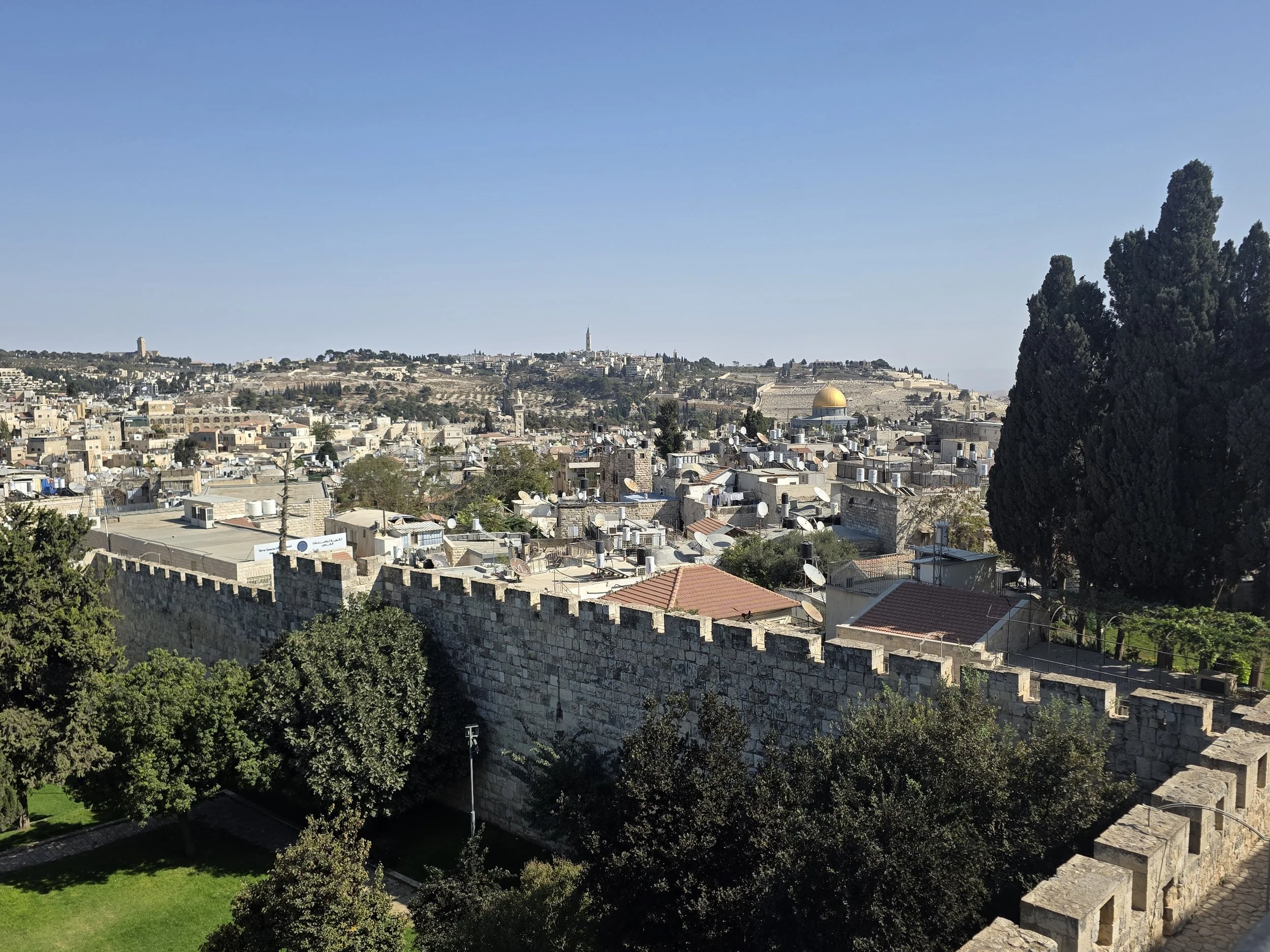 Panoramic view of Jerusalem with historic stone buildings, the golden Dome of the Rock, and surrounding hills under a clear blue sky.