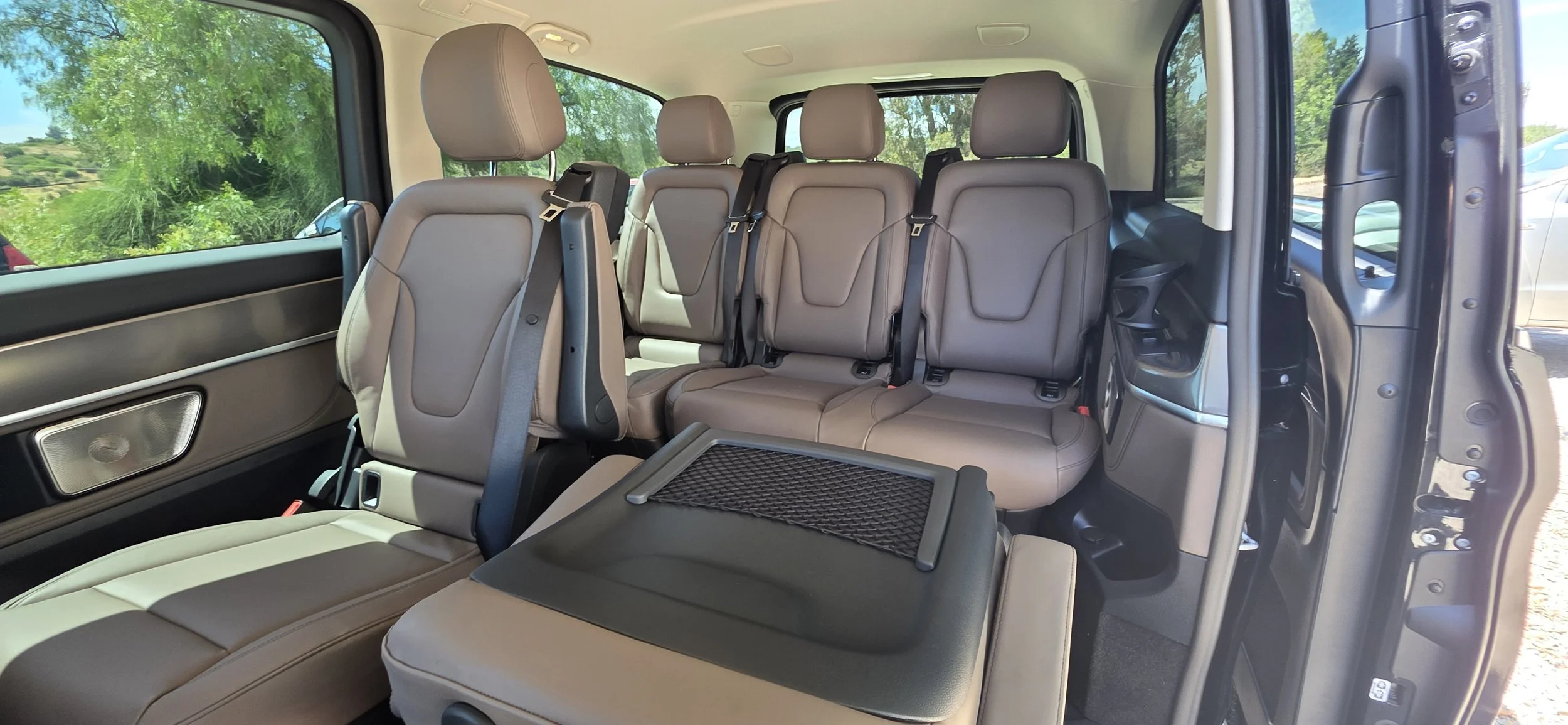 Interior of a vehicle with leather seats, showing multiple rows of seats with seat belts, and windows revealing a scenic outdoor setting with green trees.