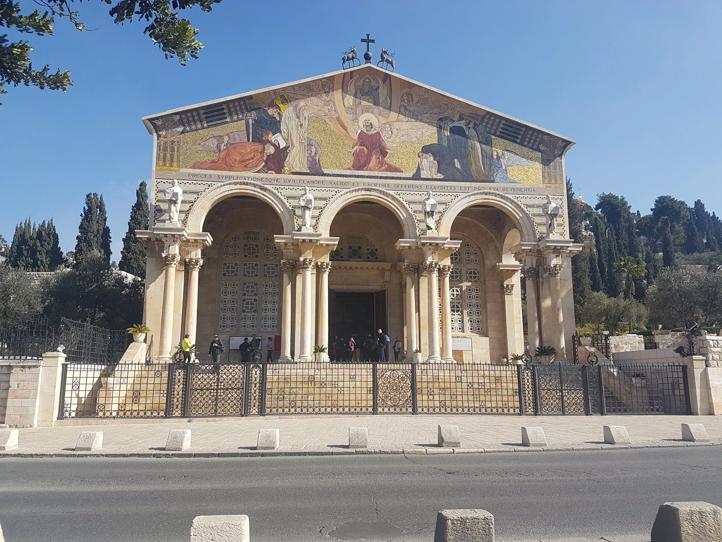 A church building with a mosaic facade depicting a religious scene with figures, a cross, and two animals on the roof. The building has three arched entryways with statues on the columns. There is a black iron fence in front, trees in the background, and a paved sidewalk and road with concrete blocks along the curb.