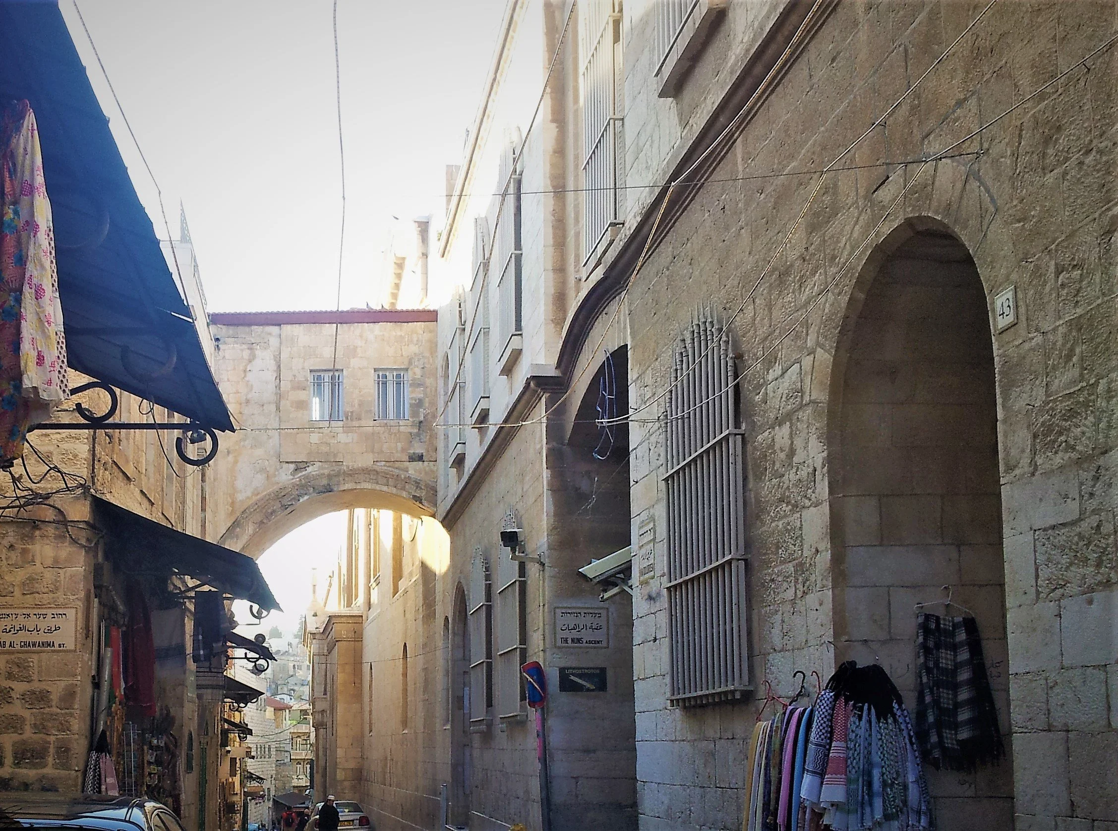 A narrow street in an urban area with stone buildings, street signs, awnings, and street vendors with colorful textiles.