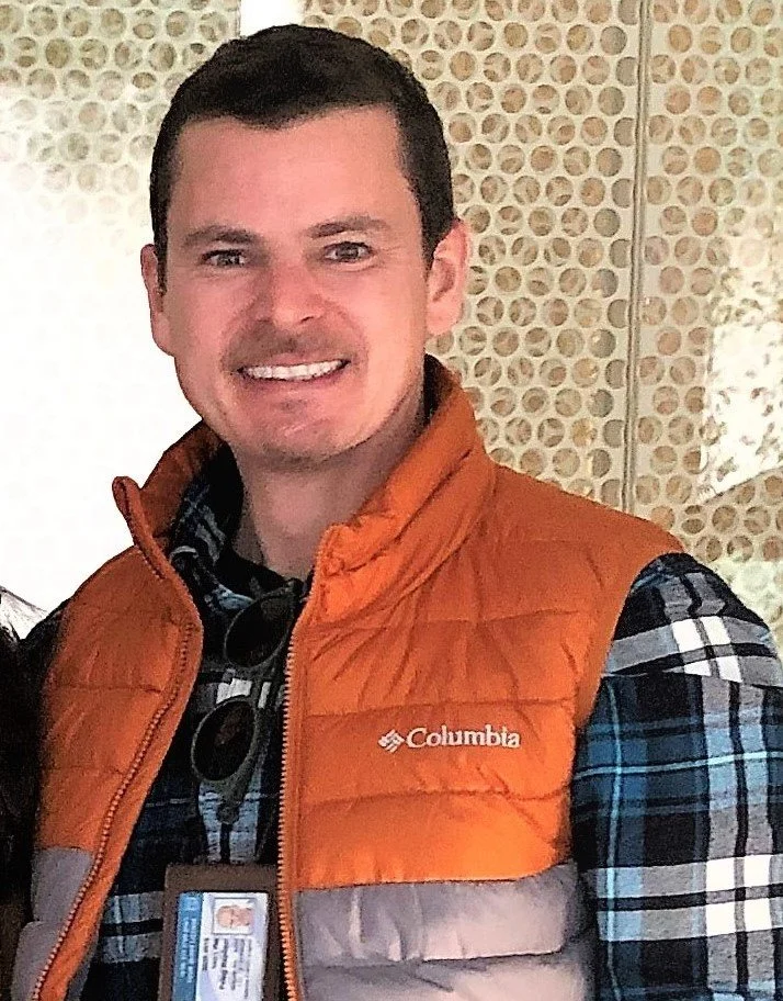 A smiling man wearing a plaid shirt, an orange Columbia puffer vest, and sunglasses hanging on his shirt. There is a patterned background behind him.