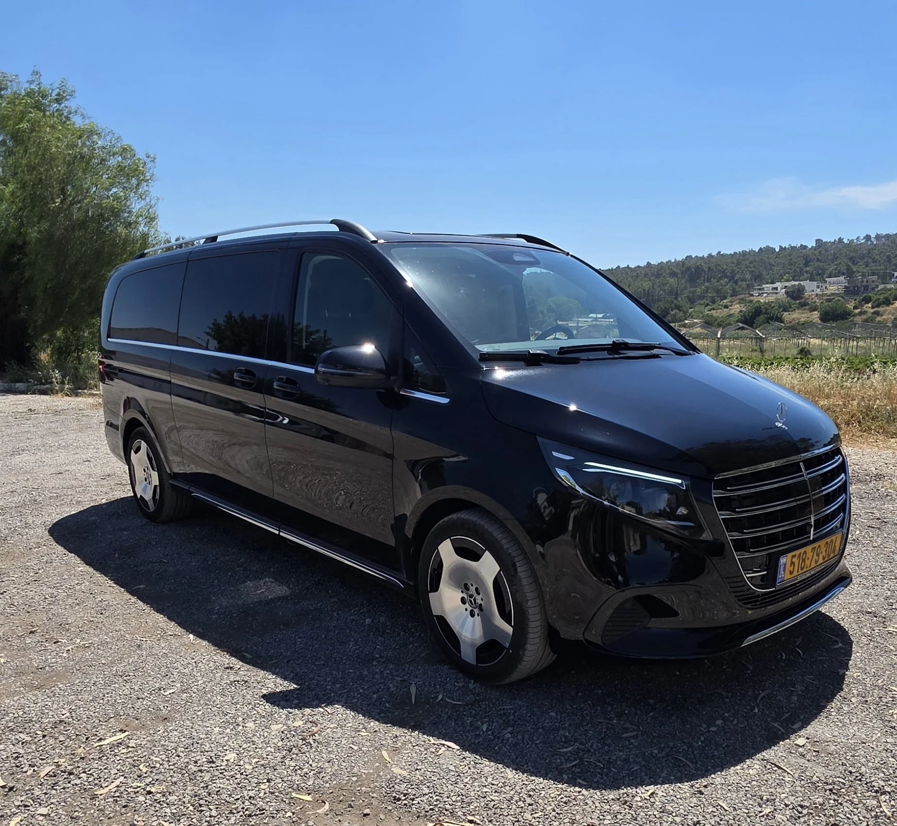 A black Mercedes-Benz van parked outdoors on a gravel surface with trees and a hillside in the background under a clear blue sky.