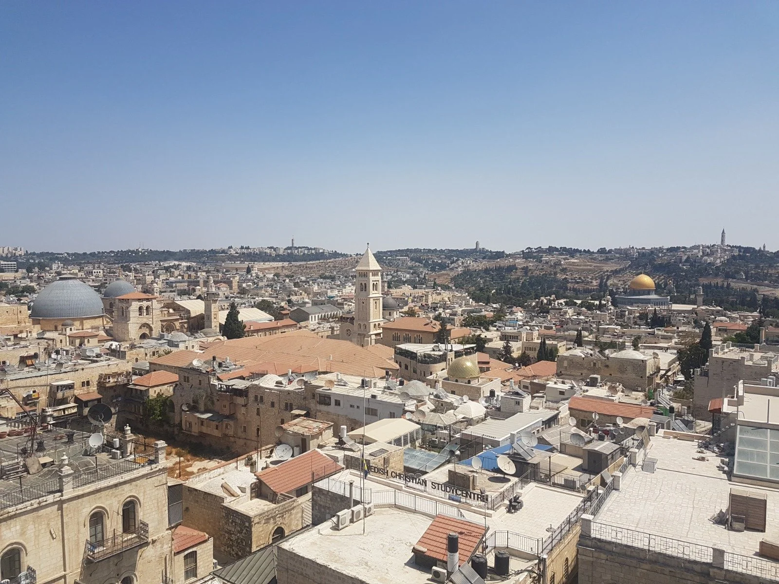 Panoramic view of Jerusalem showing historic stone buildings, domes, and church steeples under a clear blue sky.