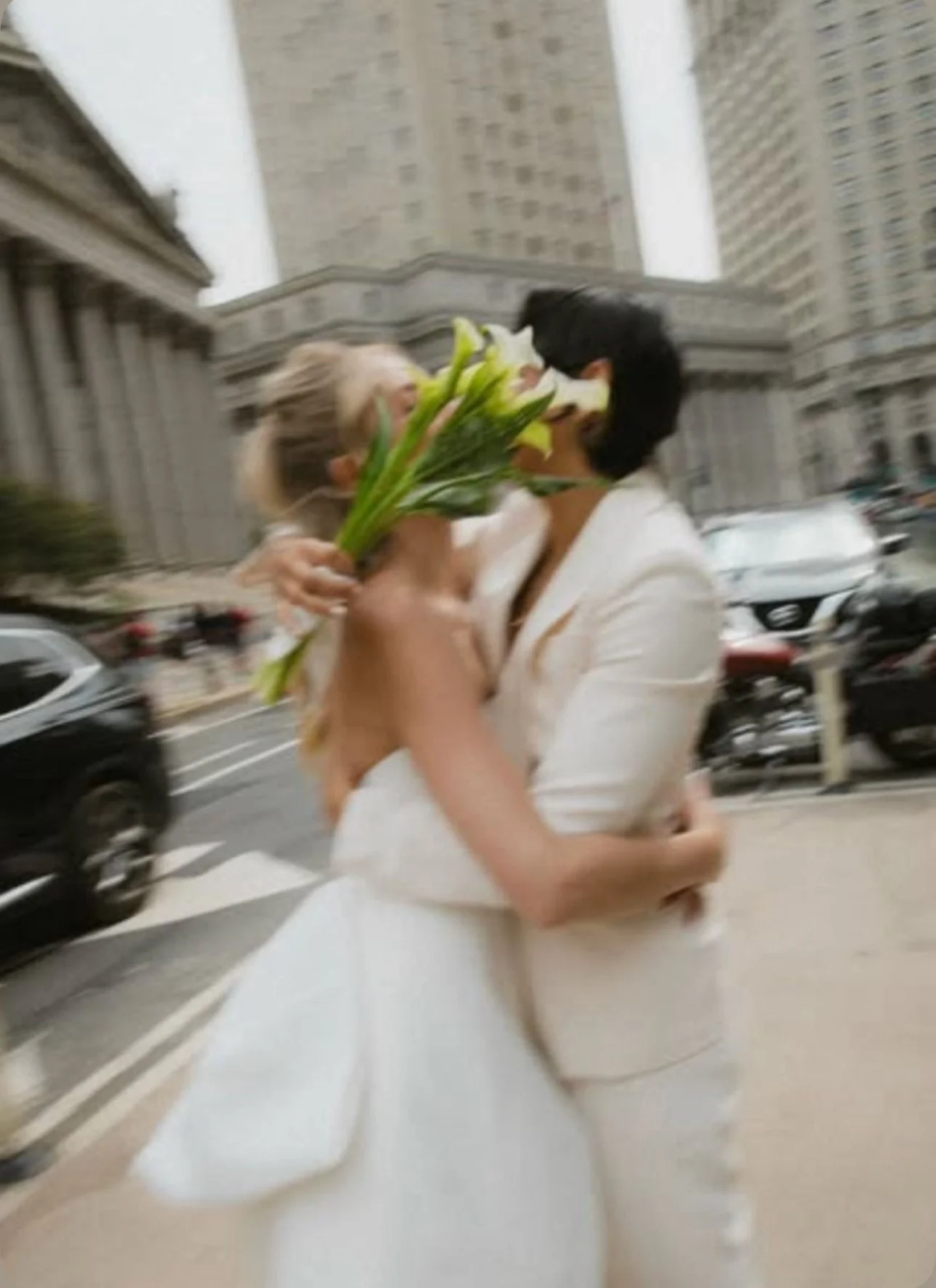 Two brides kissing on a street, with one bride wrapping her arm around the other and shielding their faces from the camera with a bouquet