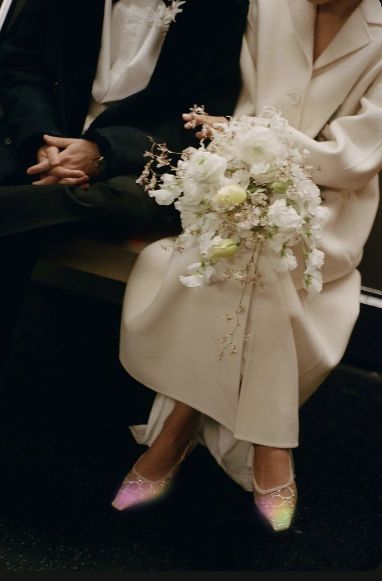 A woman wearing beige high heels with glitter, pink socks, and a beige skirt with a slit, sitting on a dark floor. She holds a bouquet of white flowers. Another person, dressed in a dark outfit with a cream blazer, is sitting beside her with their hands clasped.