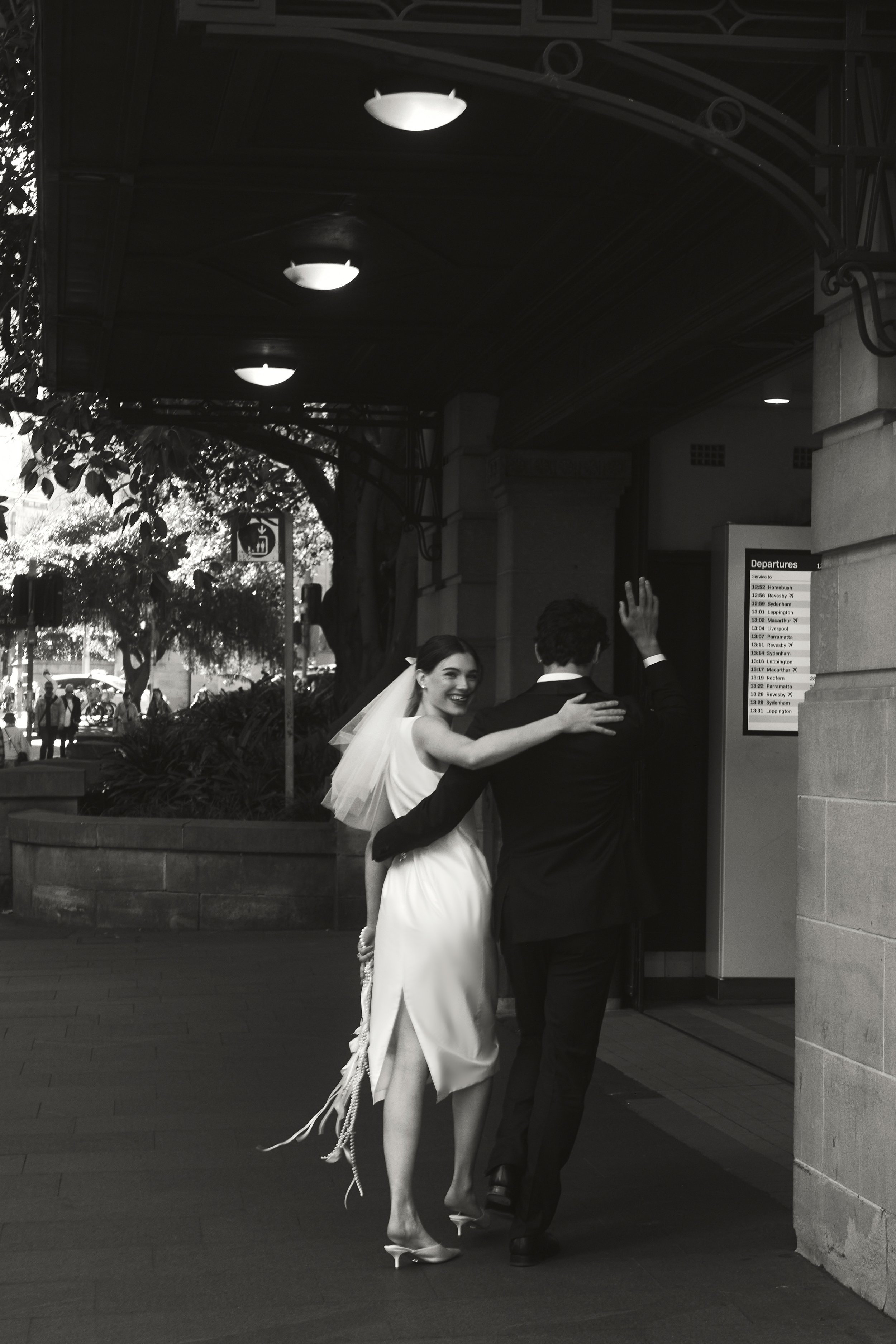 A black and white photo of a bride and groom standing outside a train station, with the bride smiling and the groom facing away. The bride is wearing a wedding dress with a veil, and the groom is in a suit. They appear happy and are possibly greeting