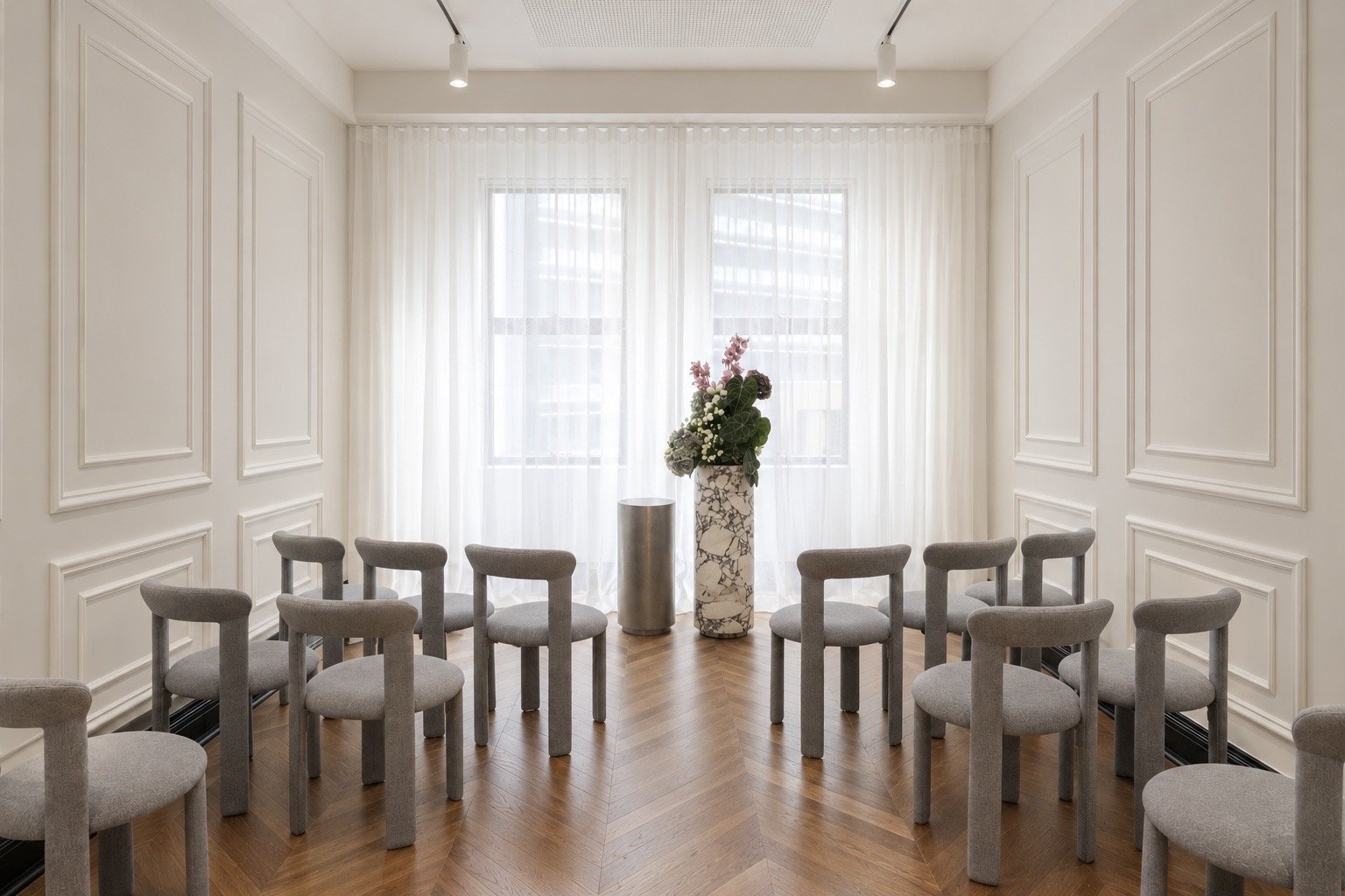 A light-drenched room with parquetry flooring, floor-to-ceiling sheer blinds, French-inspired wall paneling, marble plinths and blue upholstered chairs.