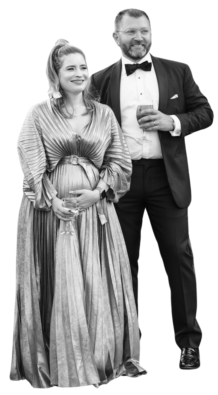 A smiling woman in a shiny, pleated gown holding a glass of wine stands next to a smiling man in a tuxedo with a bow tie, holding a glass of wine. They are at a formal event and look very happy