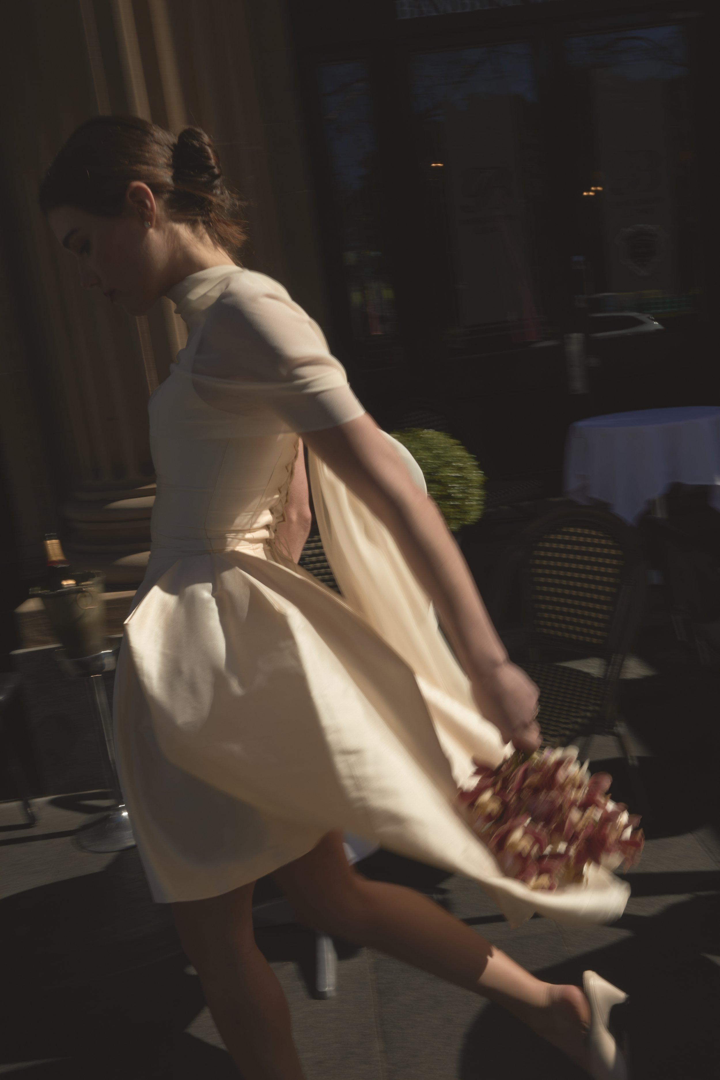 Woman in a light-colored mini bridal gown holding a bouquet of flowers, walking indoors near large windows.