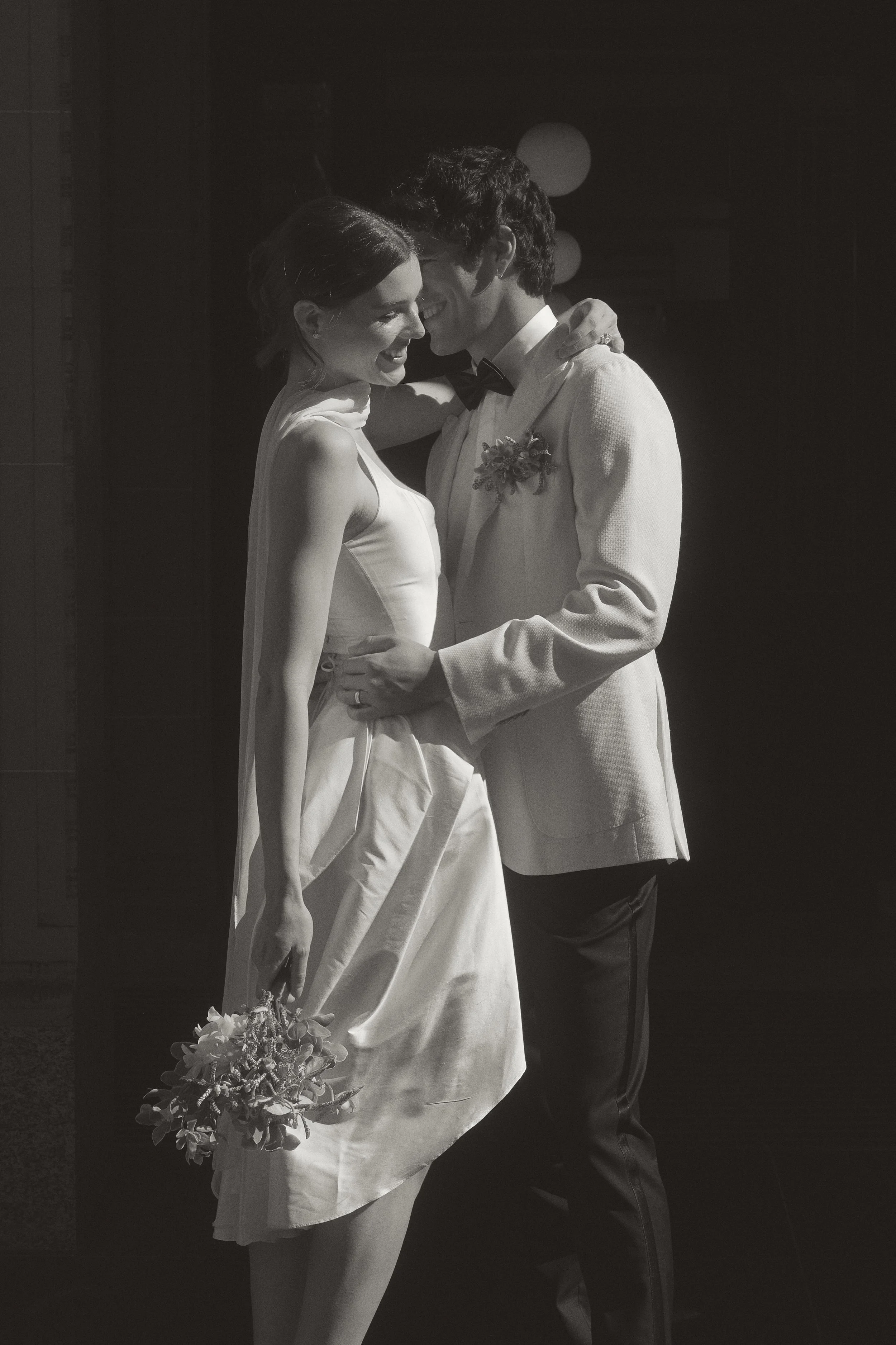 A bride and groom embrace. She carries a bouquet and his hand is on her waist.