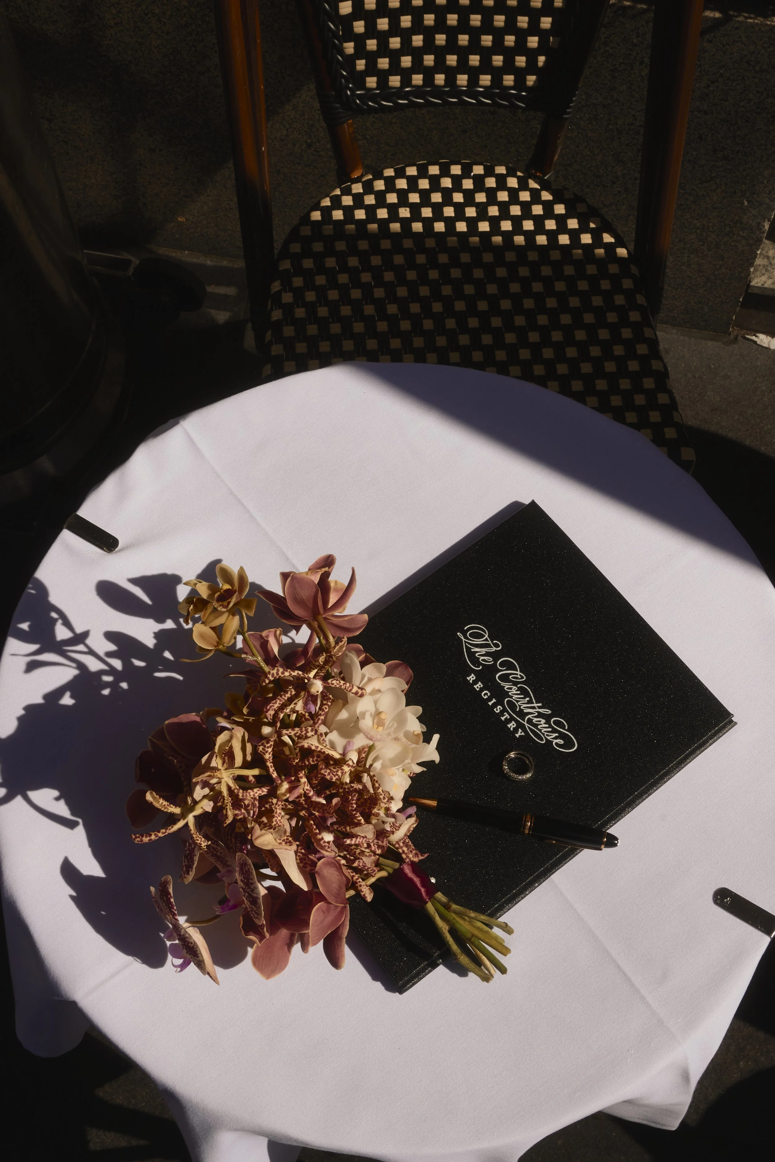 A white round table with a black menu, a pen, and a bouquet of pink orchids on it. In the background, a black and tan checked chair and a dark floor are visible.