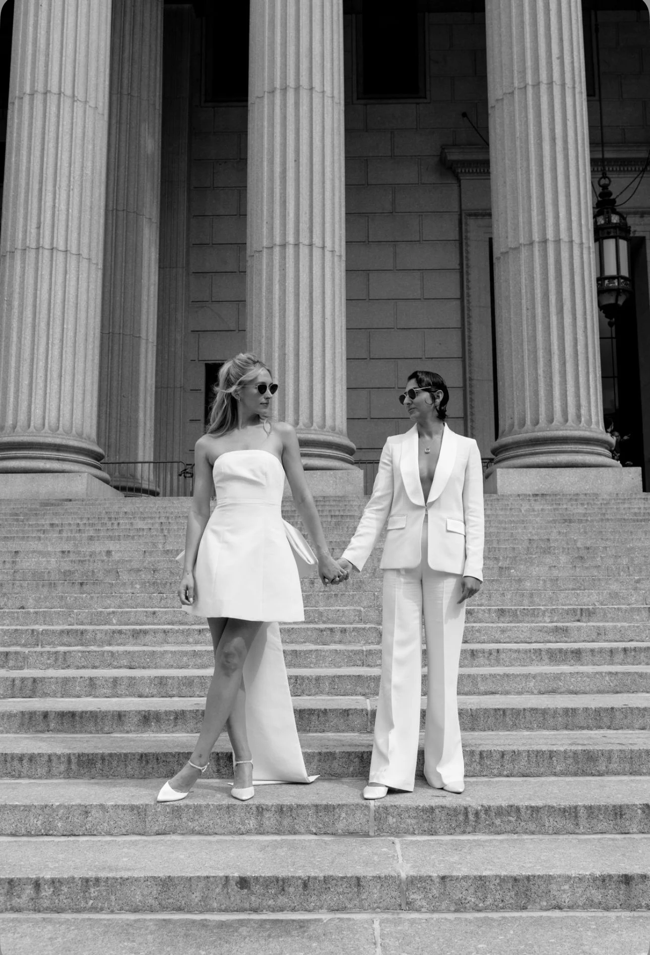 Two brides holding hands. They stand on the steps infront of large columns.