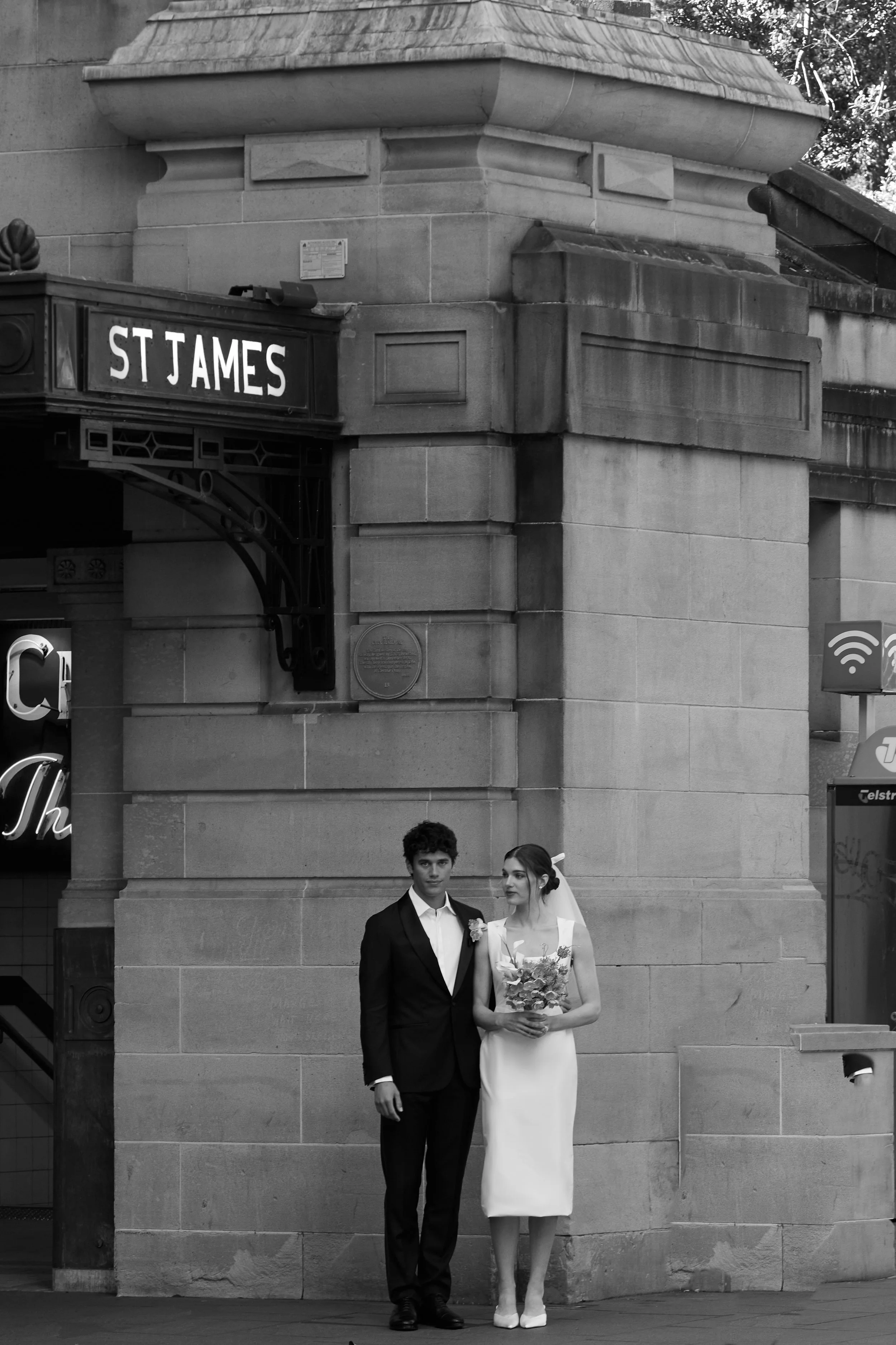 A young man in a black suit and a young woman in a white wedding dress holding a bouquet, standing together under an archway with stone columns and a sign reading 'St James' in black and white