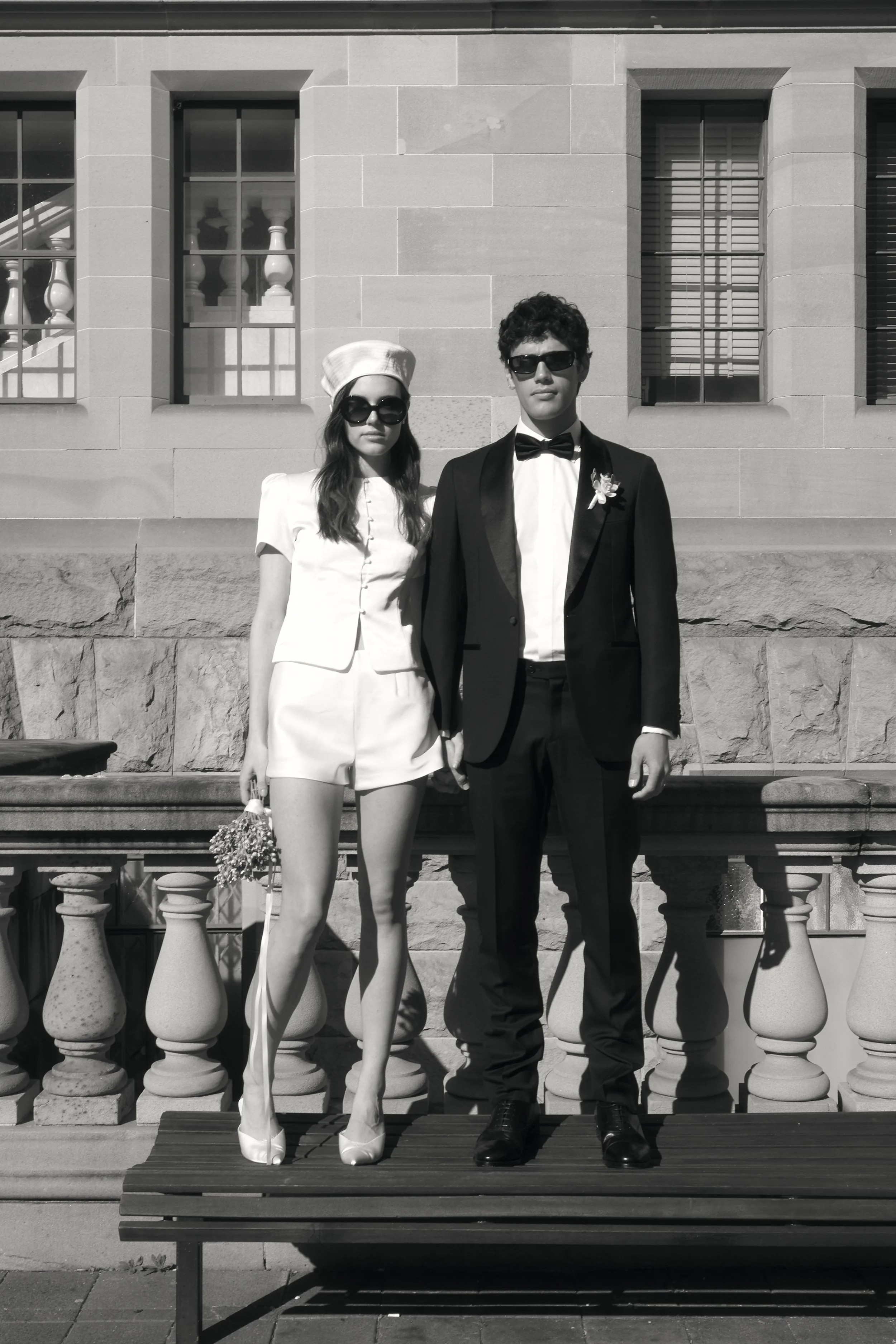 Black and white photo of a woman and a man, standing side by side on a park bench in front of a stone building with three large rectangular windows. The woman is wearing a short-sleeved dress, a hat, and large sunglasses, and is holding a small purse