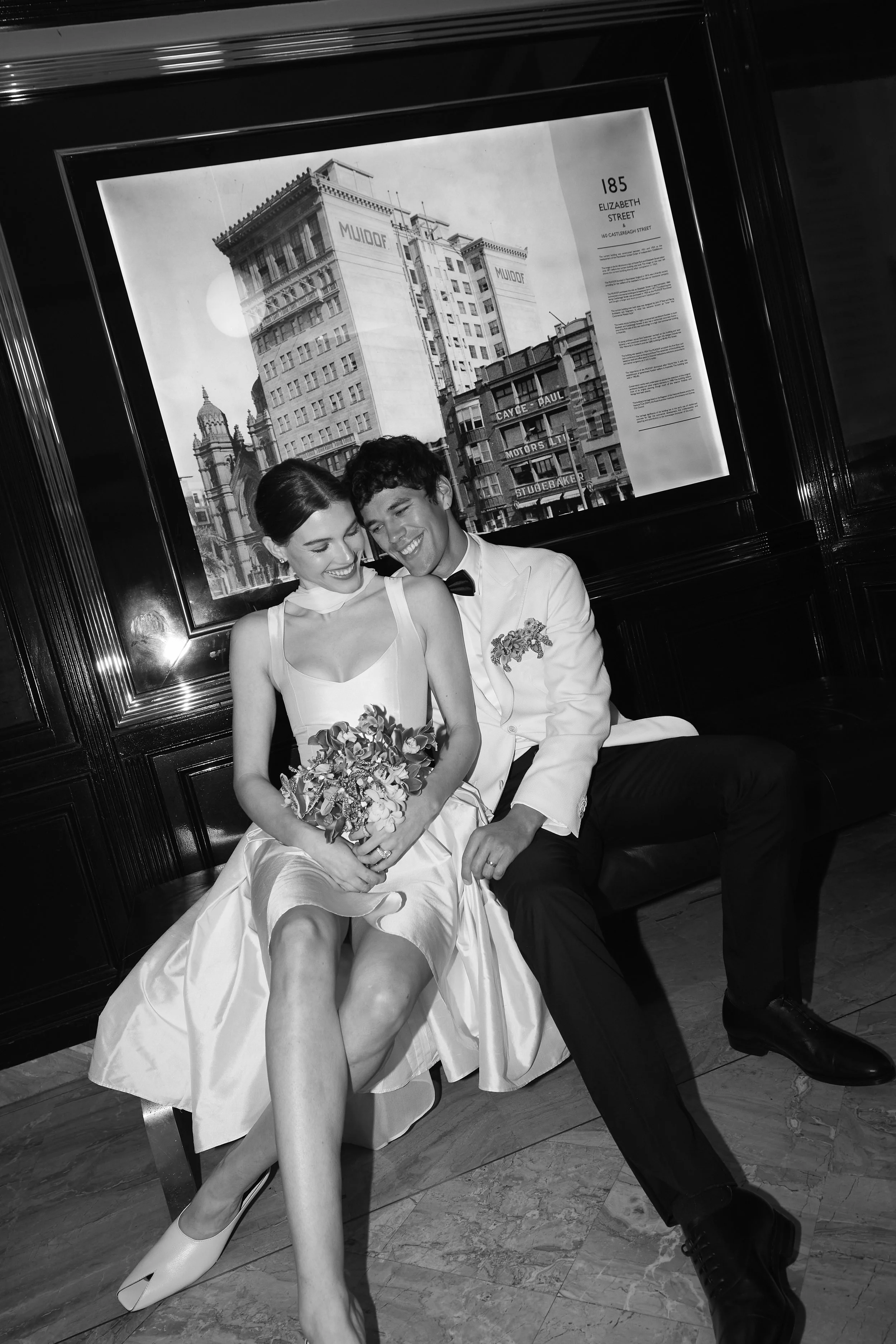 Black and white photo of a smiling woman in a wedding dress holding a bouquet and a man in a tuxedo sitting close together. They are sitting in front of a framed cityscape featuring old buildings and signage, including the address 185 Elizabeth Stree
