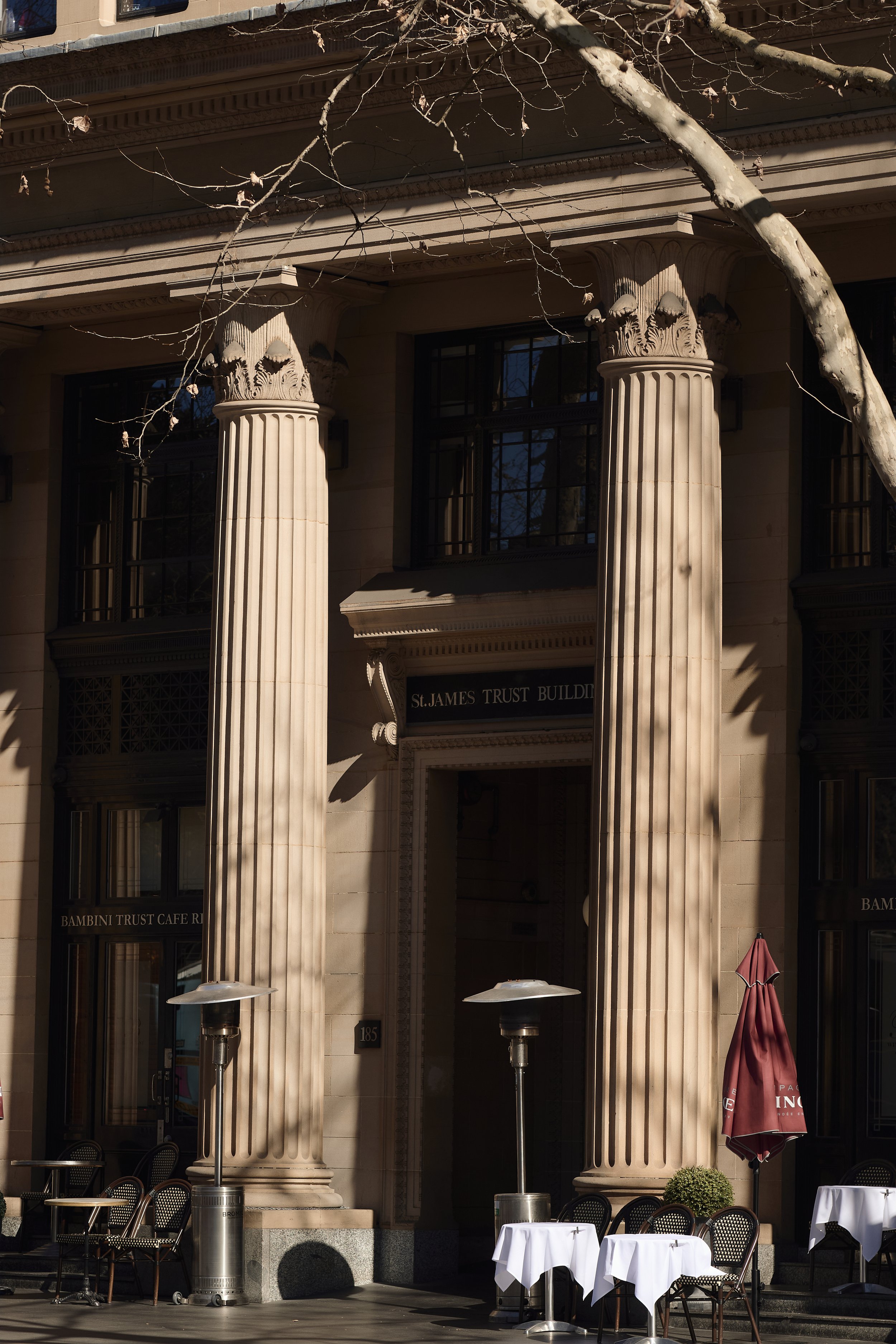 Exterior view of a building with classical architectural features such as four large, fluted columns with ornate Corinthian capitals. In front of the building, there are outdoor patio tables with white tablecloths and black chairs, as well as patio h