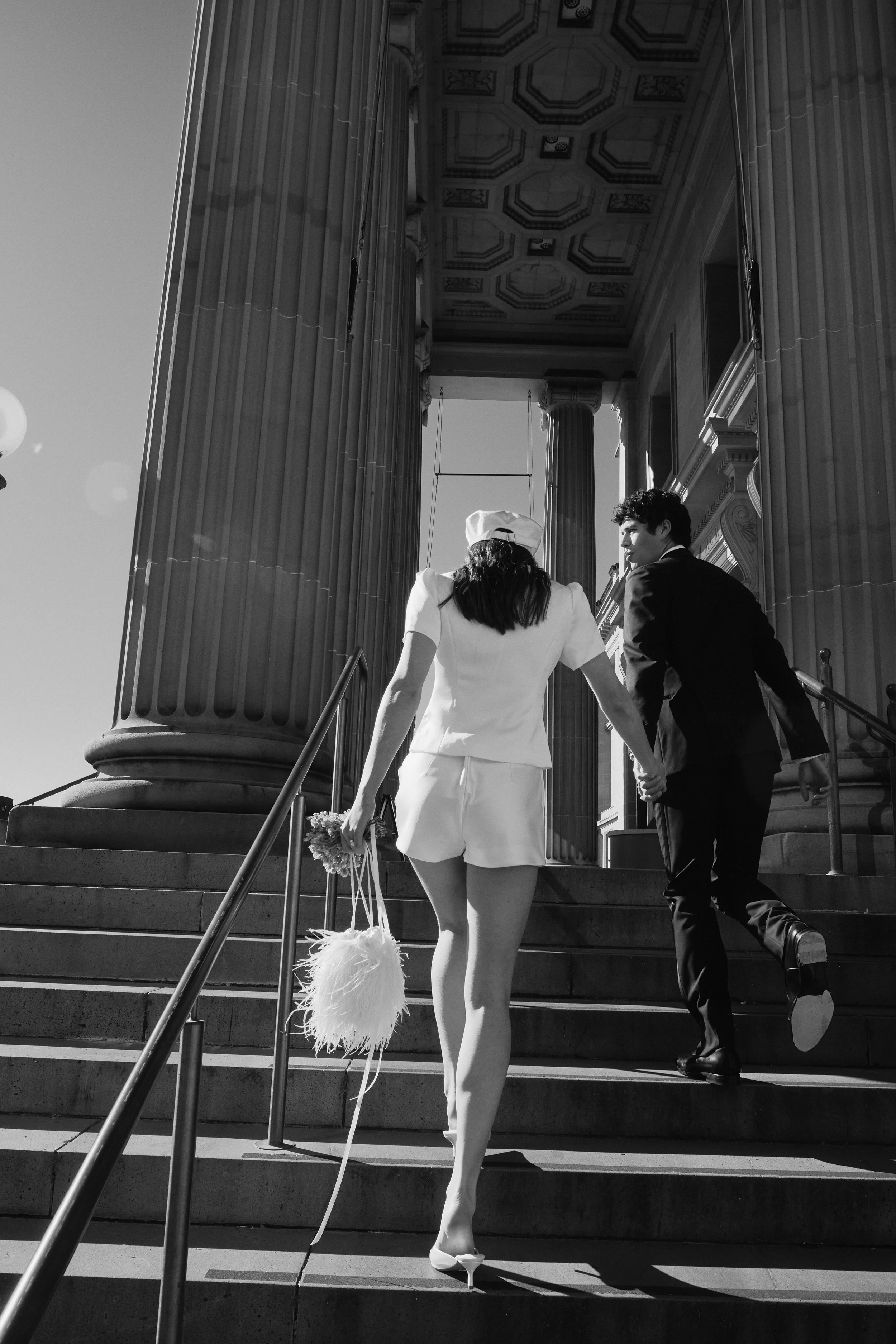 A black and white photo of a woman in a white dress and white heels holding a bouquet and a feathered clutch, and a man in a tuxedo holding hands, ascending stairs outside a grand building with large columns and detailed ceiling