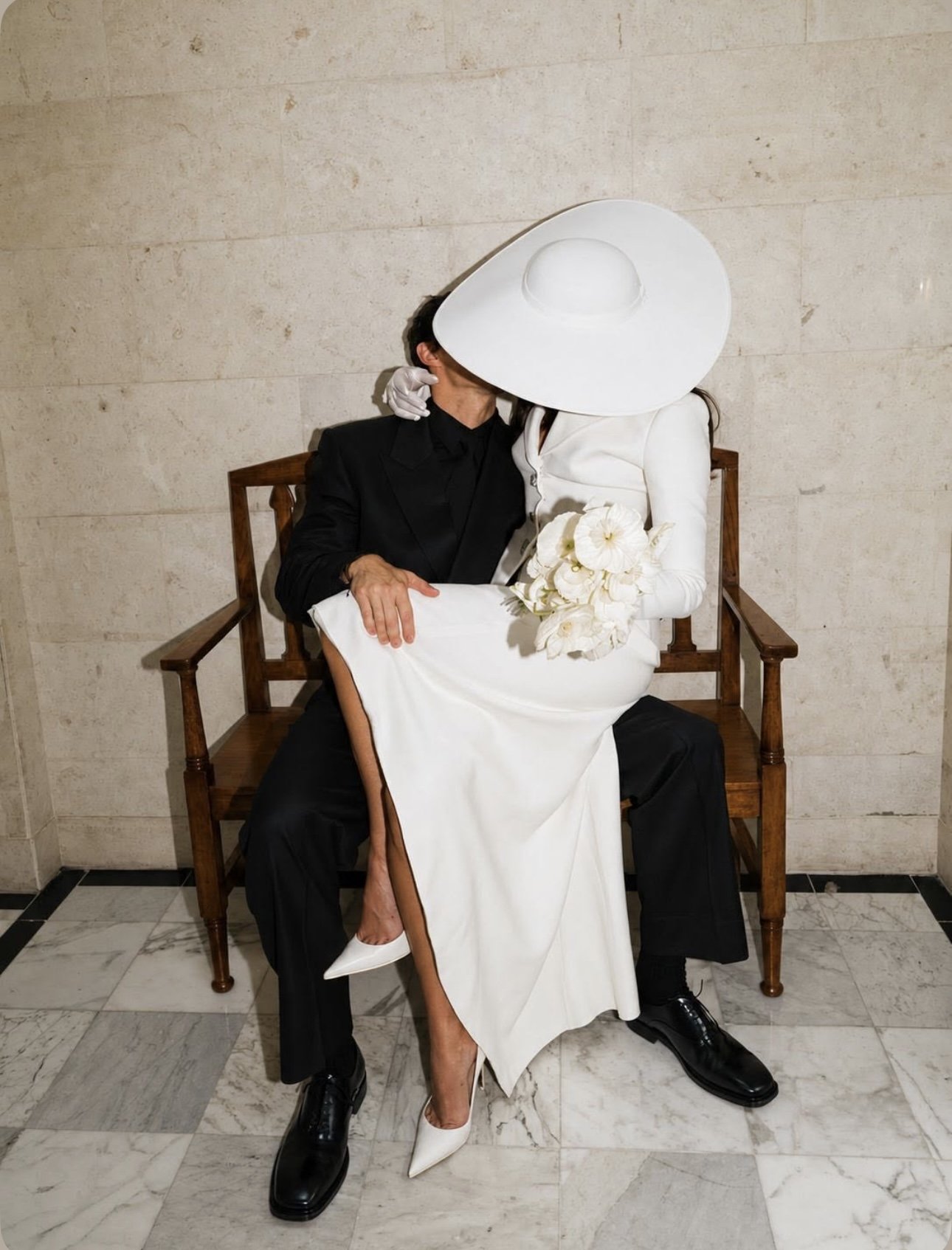 A couple sitting on a wooden bench, embracing and kissing. The woman is dressed in a white gown, large white hat, and holding white flowers. The man is dressed in black, wearing a suit and shoes. The couple are kissing and their faces are shielded