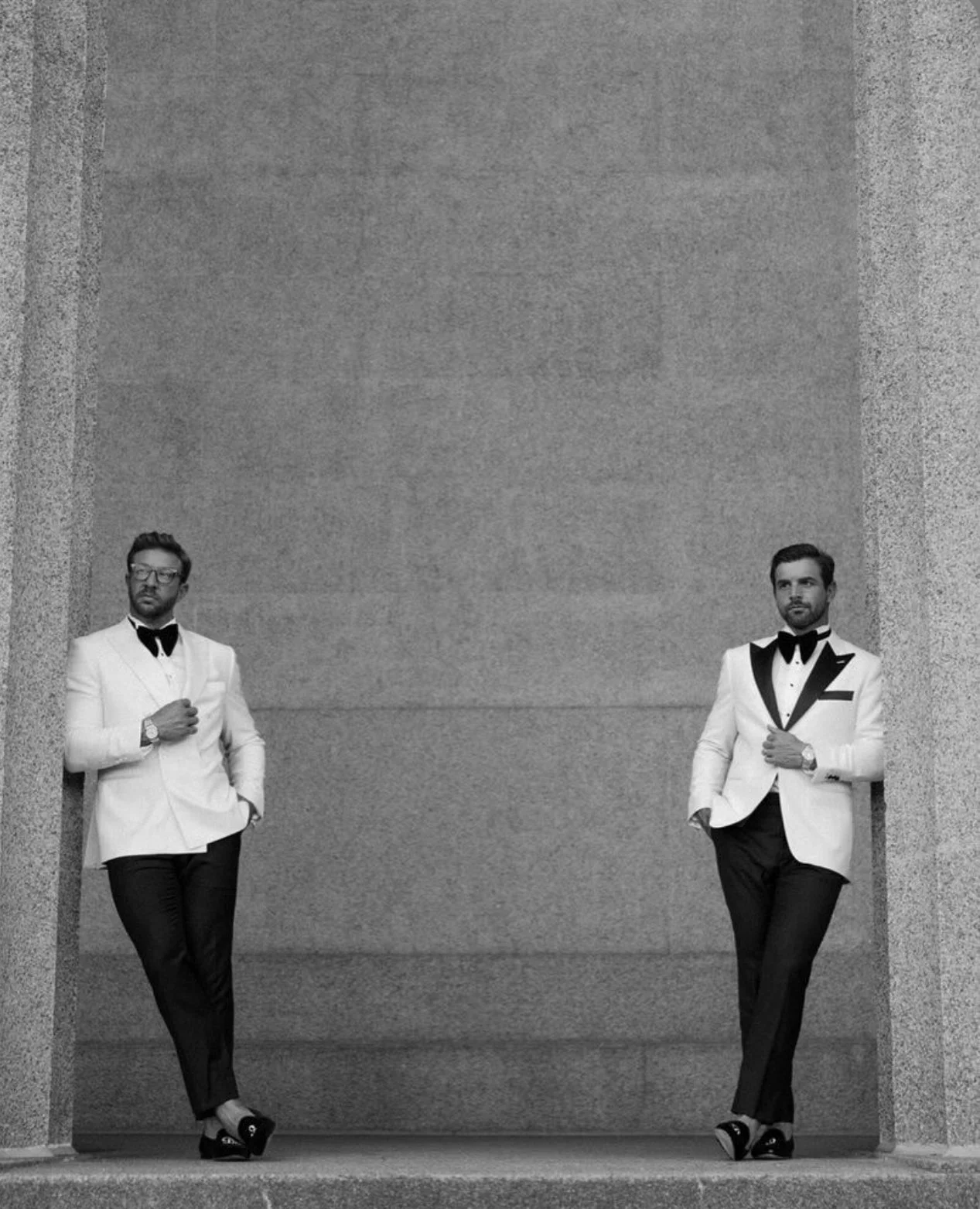 Two grooms in white tuxedo suits with bow ties, standing in a corridor with textured walls, one on each side, looking in opposite directions.