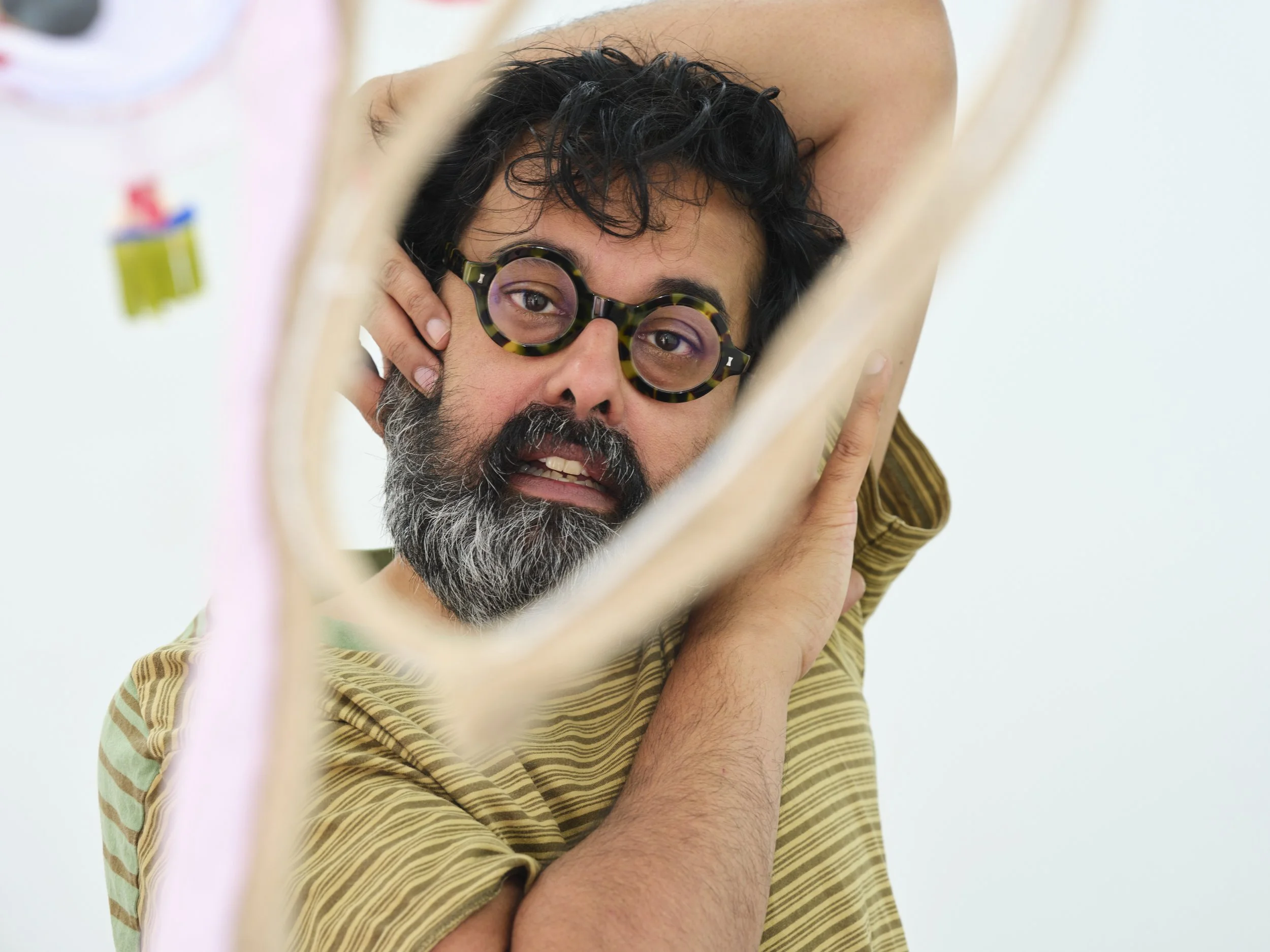 A man with dark, curly hair and a beard is lying down, looking into a mirror, wearing tortoiseshell glasses, with a woman behind him gently holding his face.