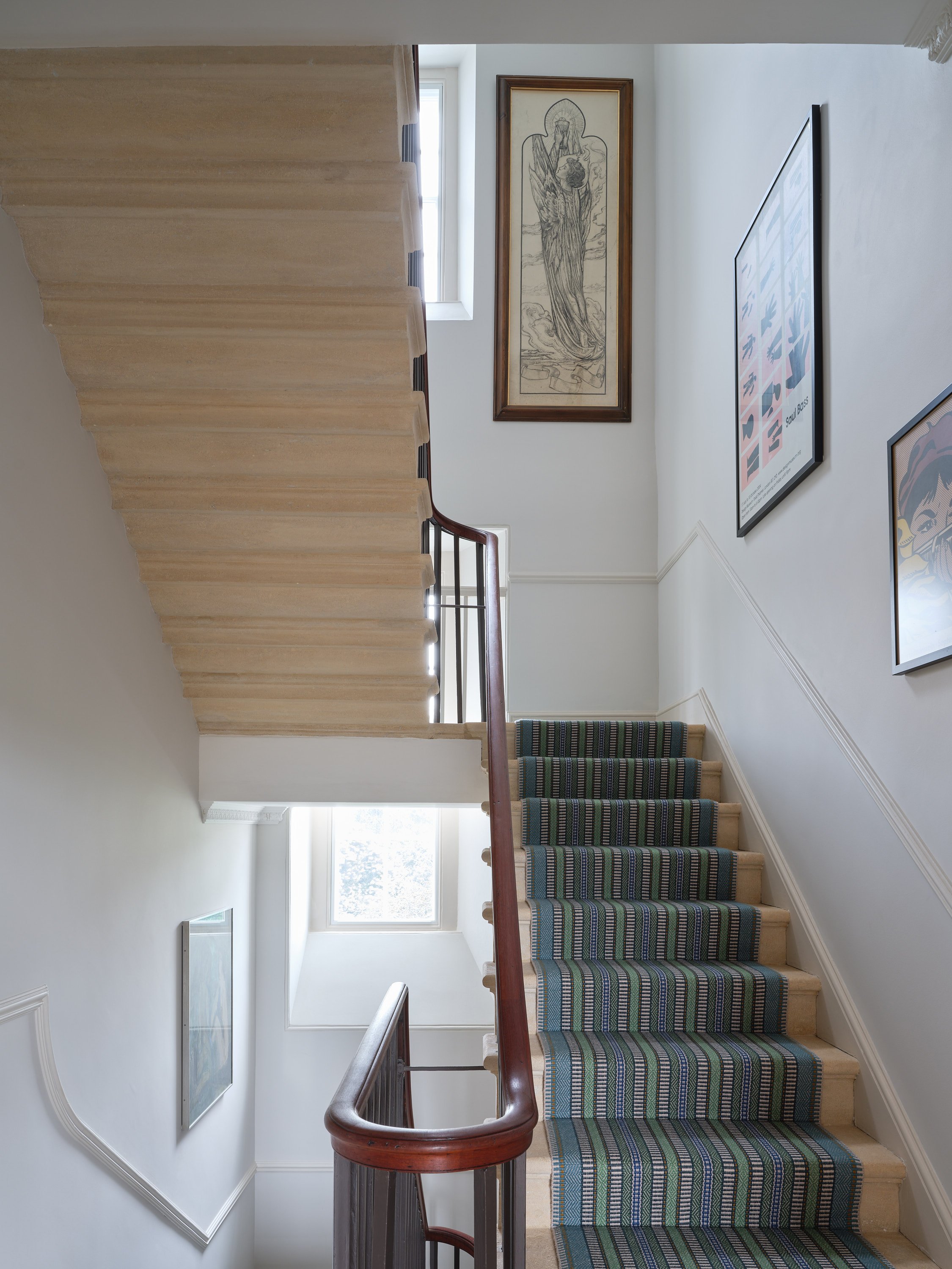 A staircase with a striped green and blue carpet runner, a wooden banister, and white walls decorated with framed artwork, including a large portrait and a smaller illustration. Natural light filters through windows at the top of the stairs.