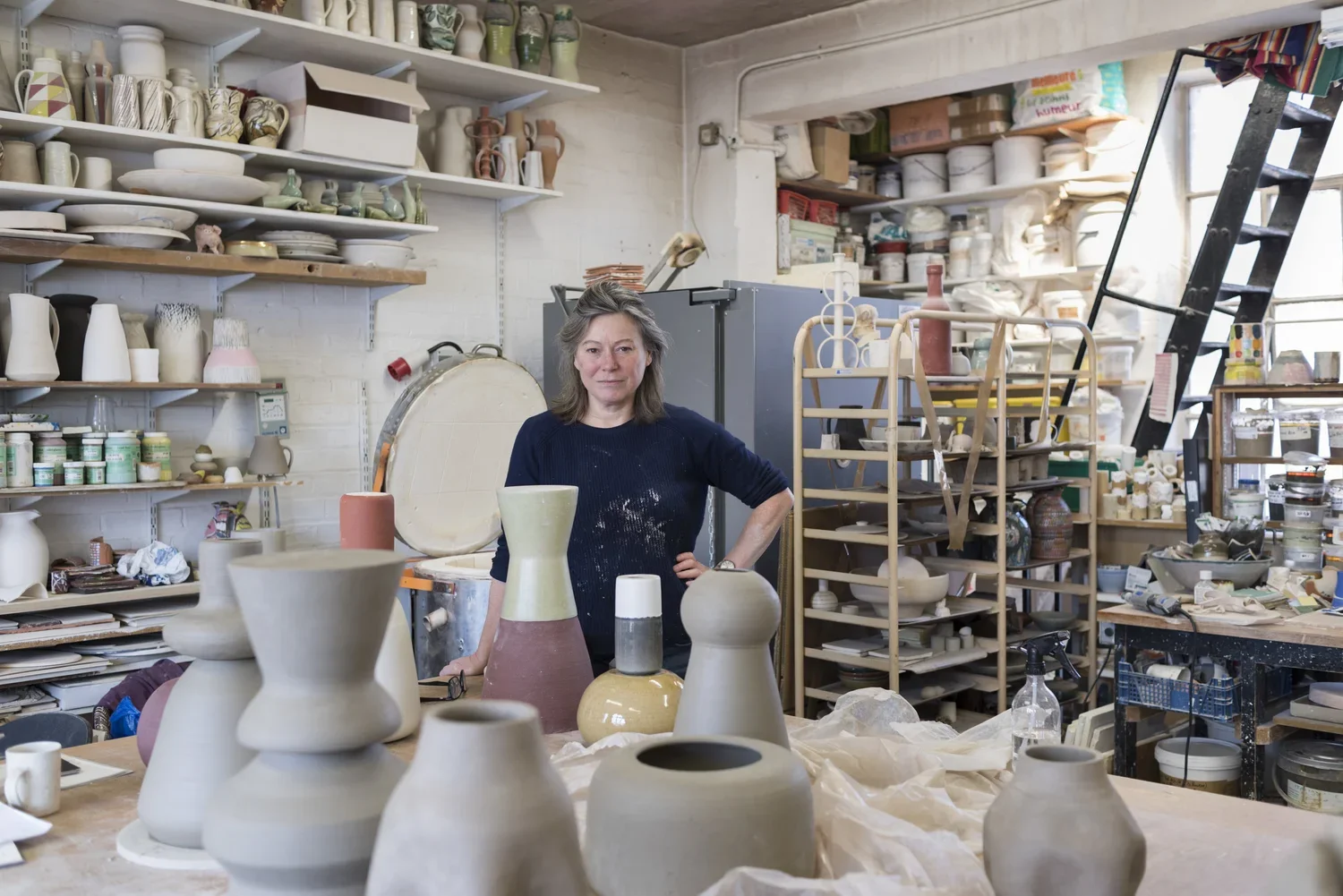 A woman standing in a pottery studio surrounded by various ceramic vases and pottery pieces in progress, shelves filled with pottery supplies, and a ladder near a window.