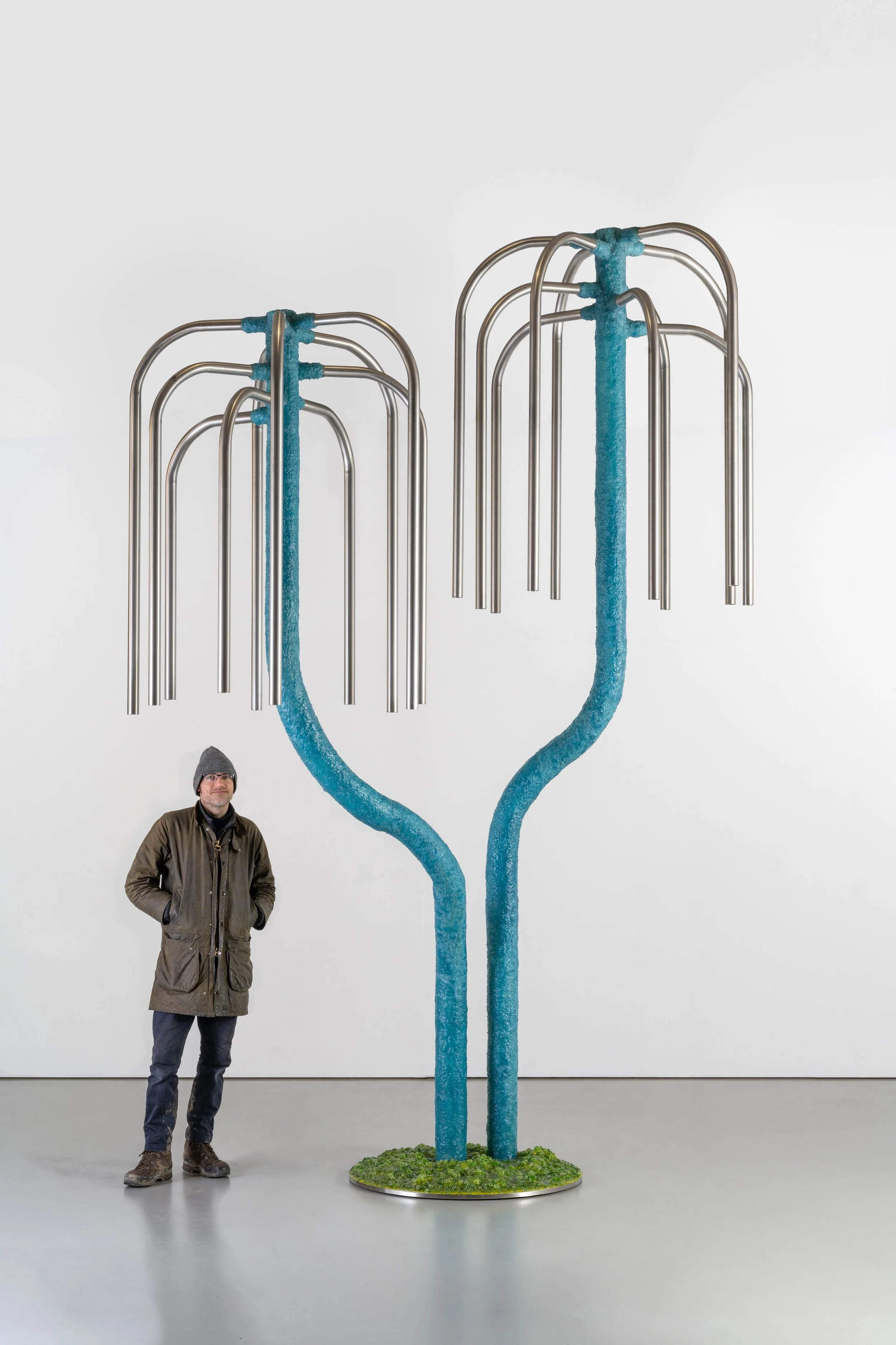 A man stands in front of an art installation resembling two large drinking straws with sets of metal bendy tubes, all painted with a textured blue coating, set against a plain white wall.