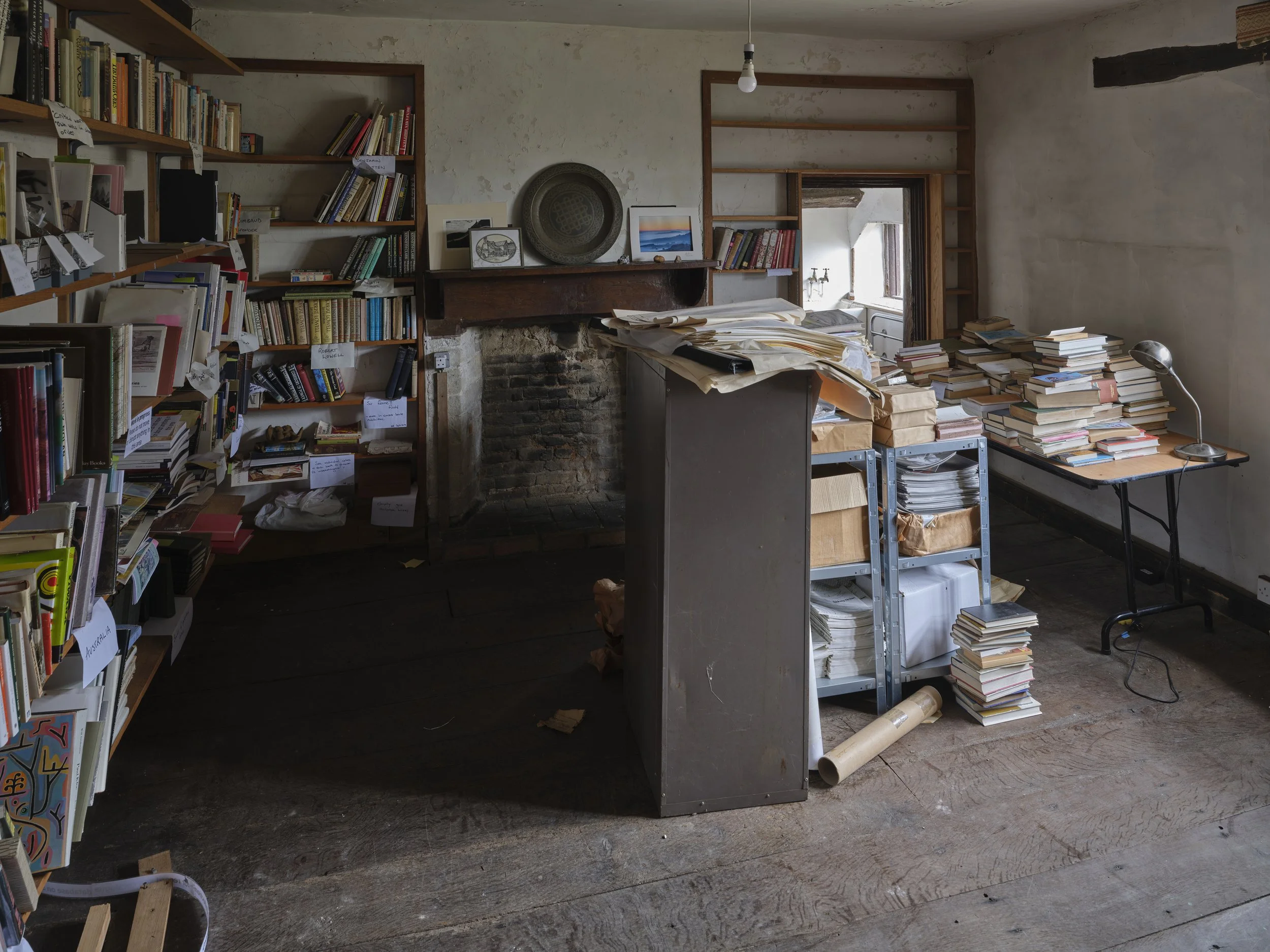 Disorganized room with bookshelves, stacks of books, papers, and miscellaneous items in an old house with a brick fireplace.